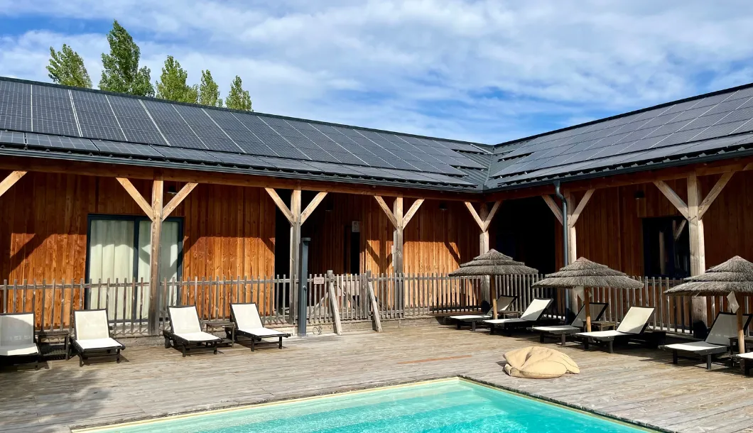 Outdoor pool with wooden deck, lounge chairs, umbrellas, and wooden building with solar panels on roof.