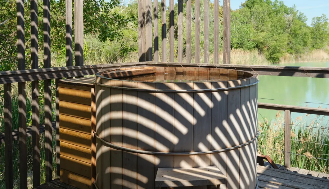 Wooden hot tub on a deck with shadows, surrounded by green trees and a body of water.