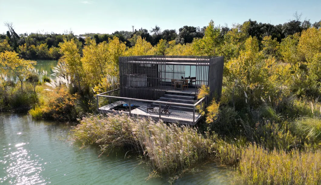 Modern wooden cottage with deck by a lake surrounded by autumn trees and reeds in bright sunlight.