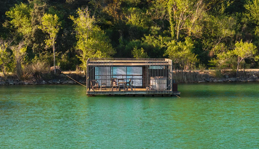 Modern wooden floating cabin on a green lake with forest in the background.