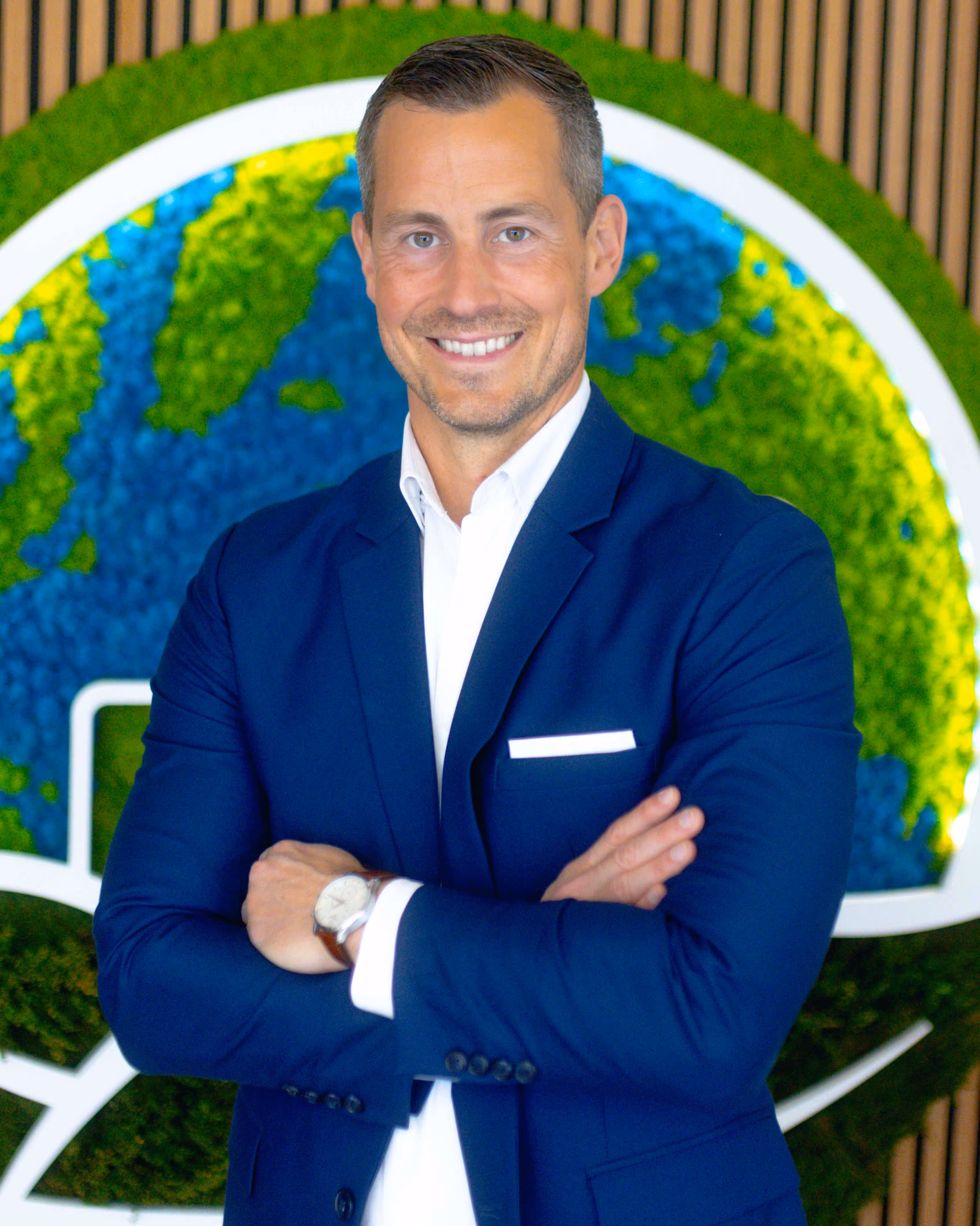 Smiling man in a blue suit with arms crossed standing in front of a green and blue earth-themed wall decoration.