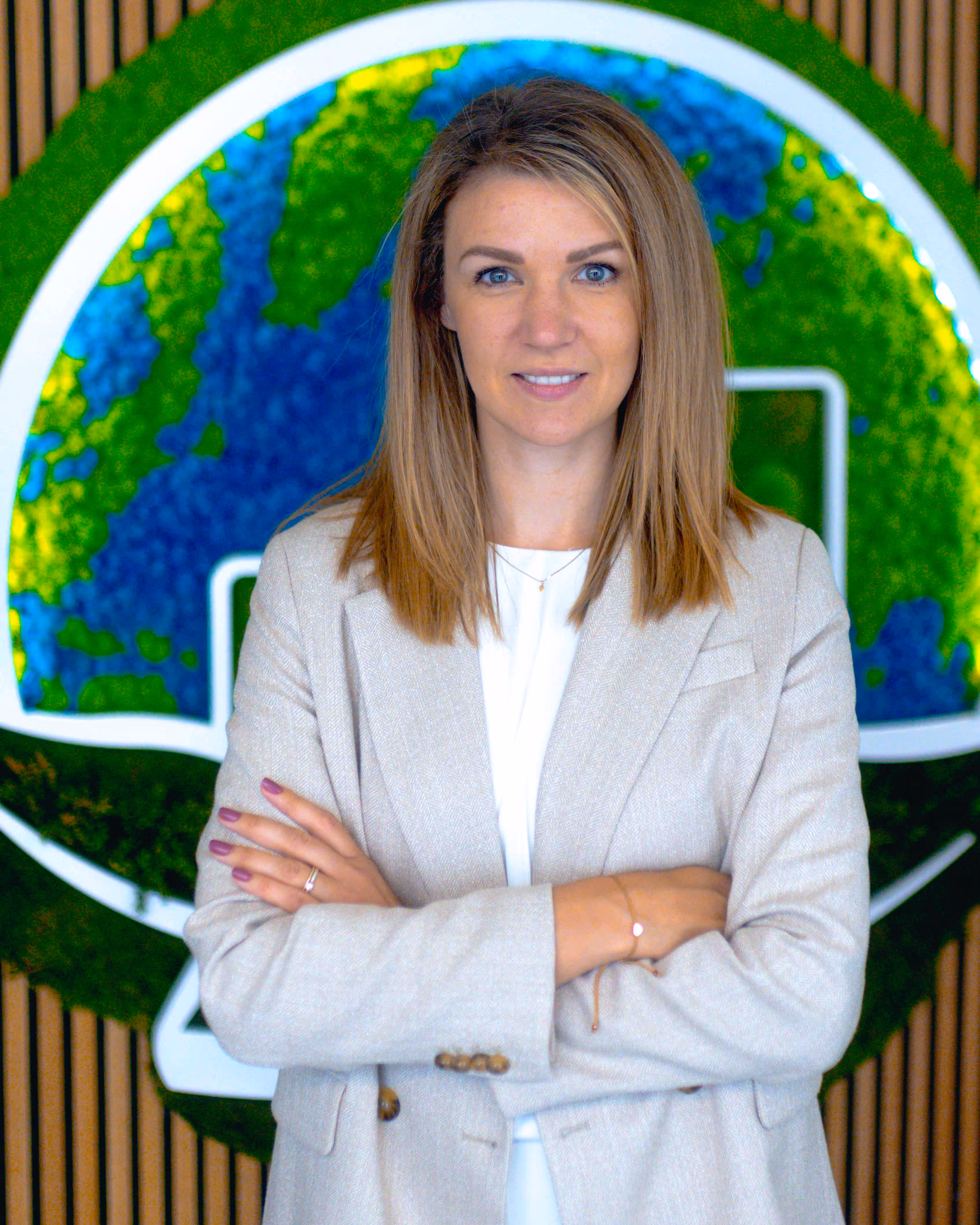 Woman with shoulder-length blonde hair wearing a light beige blazer, standing with arms crossed in front of a green and blue circular logo on a wooden slat wall.
