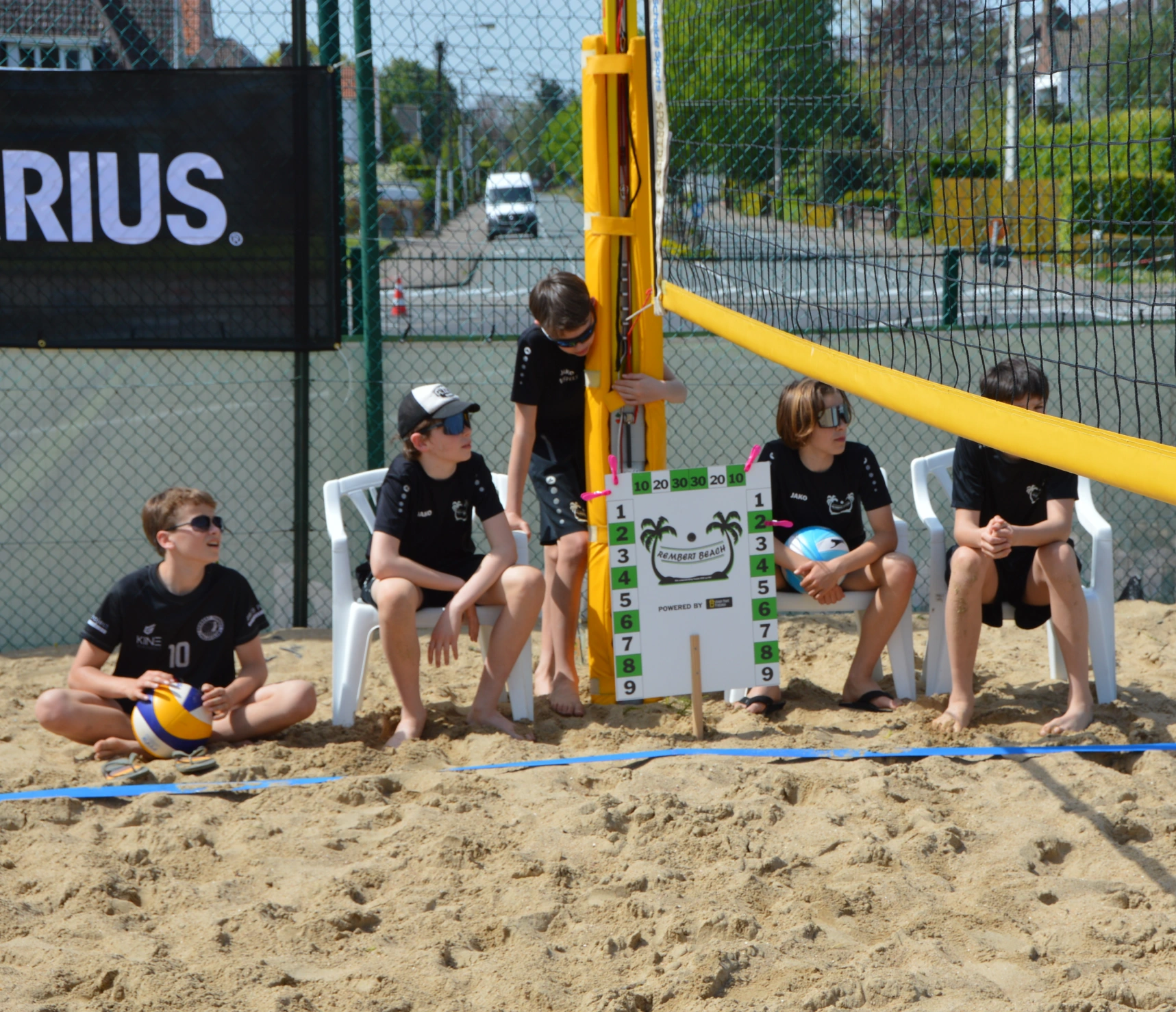 Vier jongens zitten in het zand naast een beachvolleybalveld; één buigt over een scorebord met het Rembert Beach-logo tijdens een beachvolleybalwedstrijd.