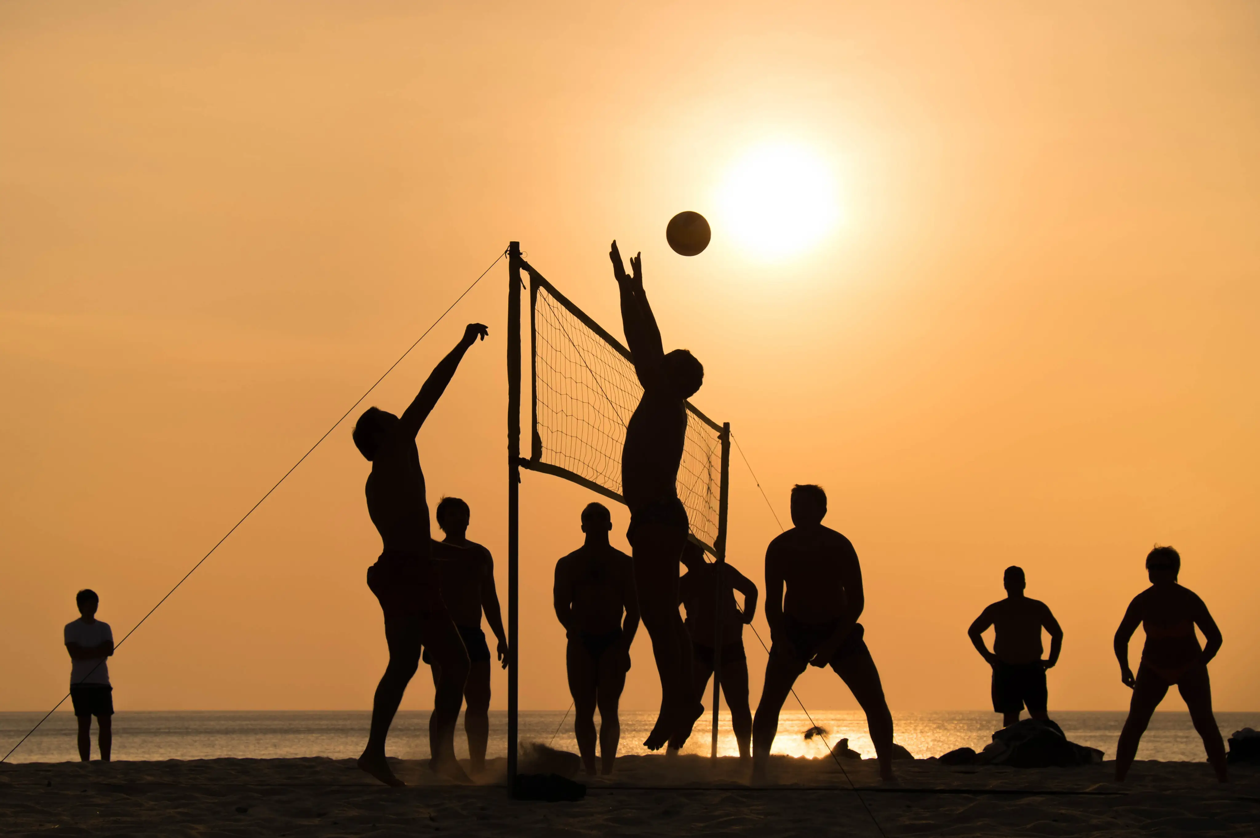 Enkele mensen spelen beachvolleybal op het strand met op de achtergrond de zee en de fel schijnende zon.