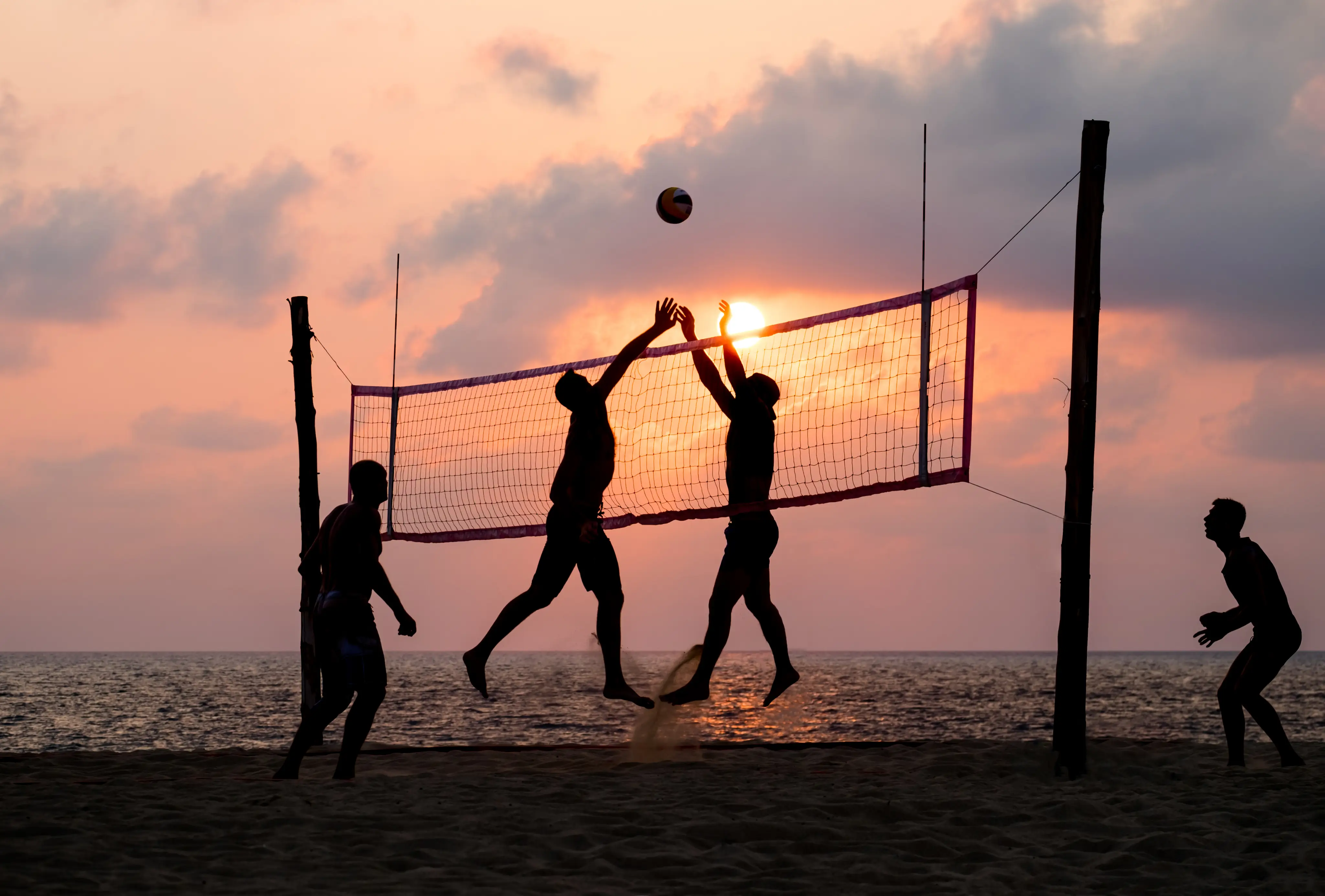 Enkele mensen spelen beachvolleybal op het strand met op de achtergrond de zee en de fel schijnende zon.
