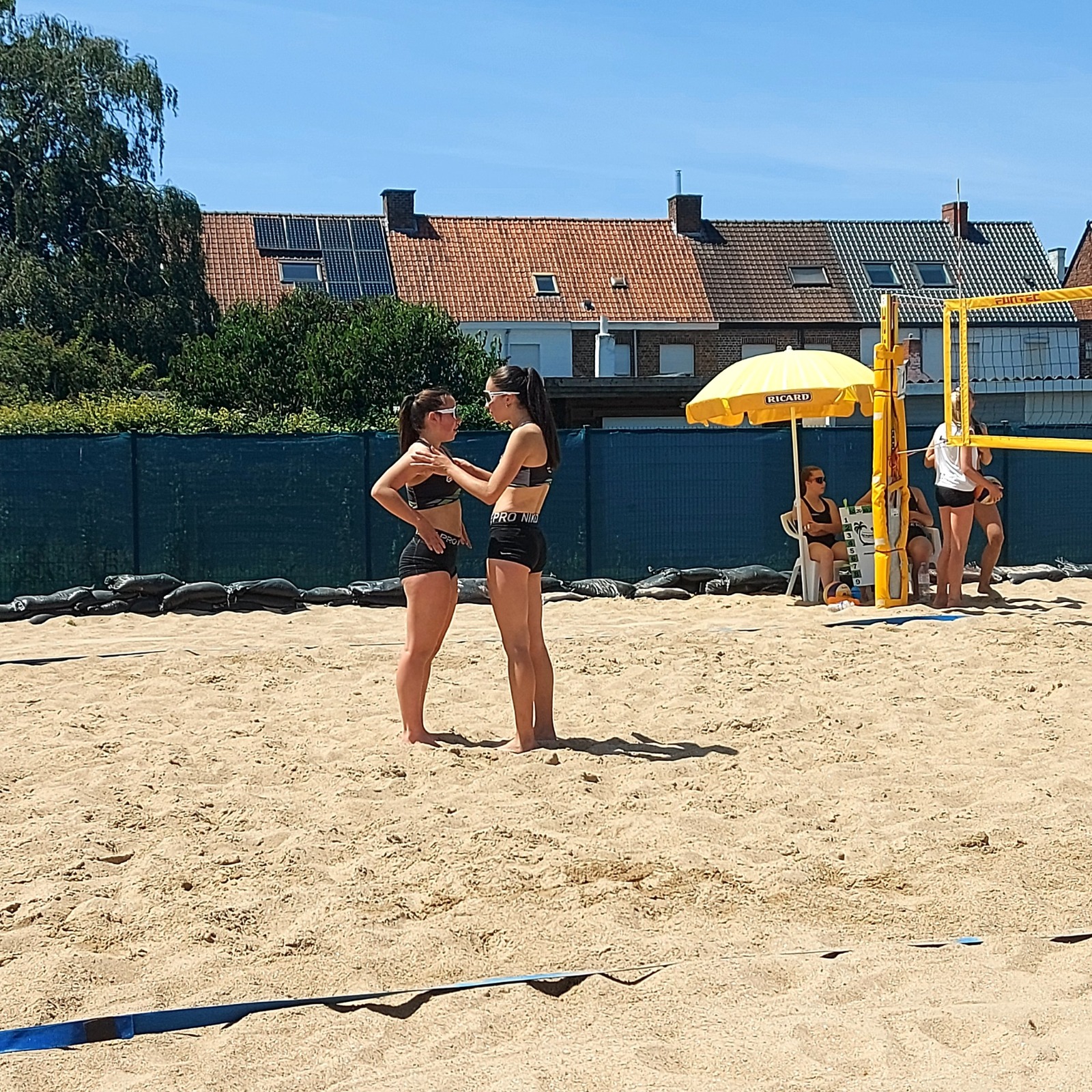 Twee vrouwelijke beachvolleybalspelers in zwarte sportkleding overleggen op het zand met een volleybalnet en toeschouwers met gele parasol op de achtergrond.