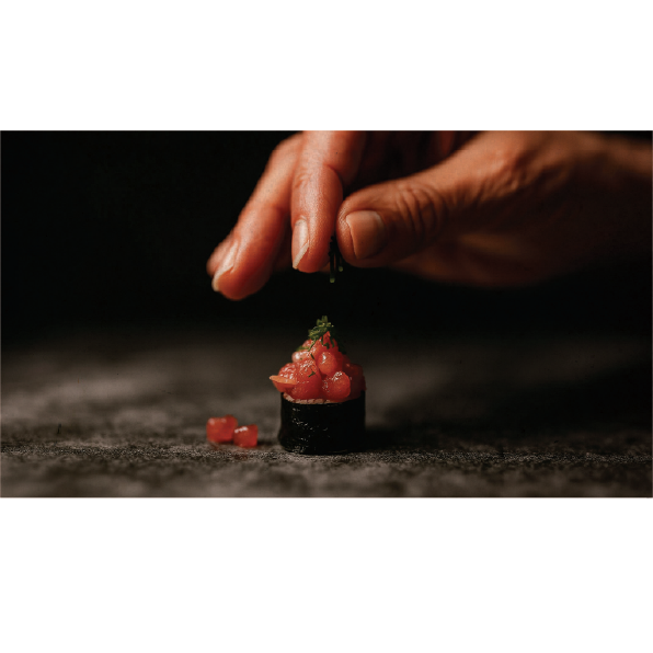 Hand sprinkling green garnish over small sushi roll topped with diced raw fish on a dark textured surface.