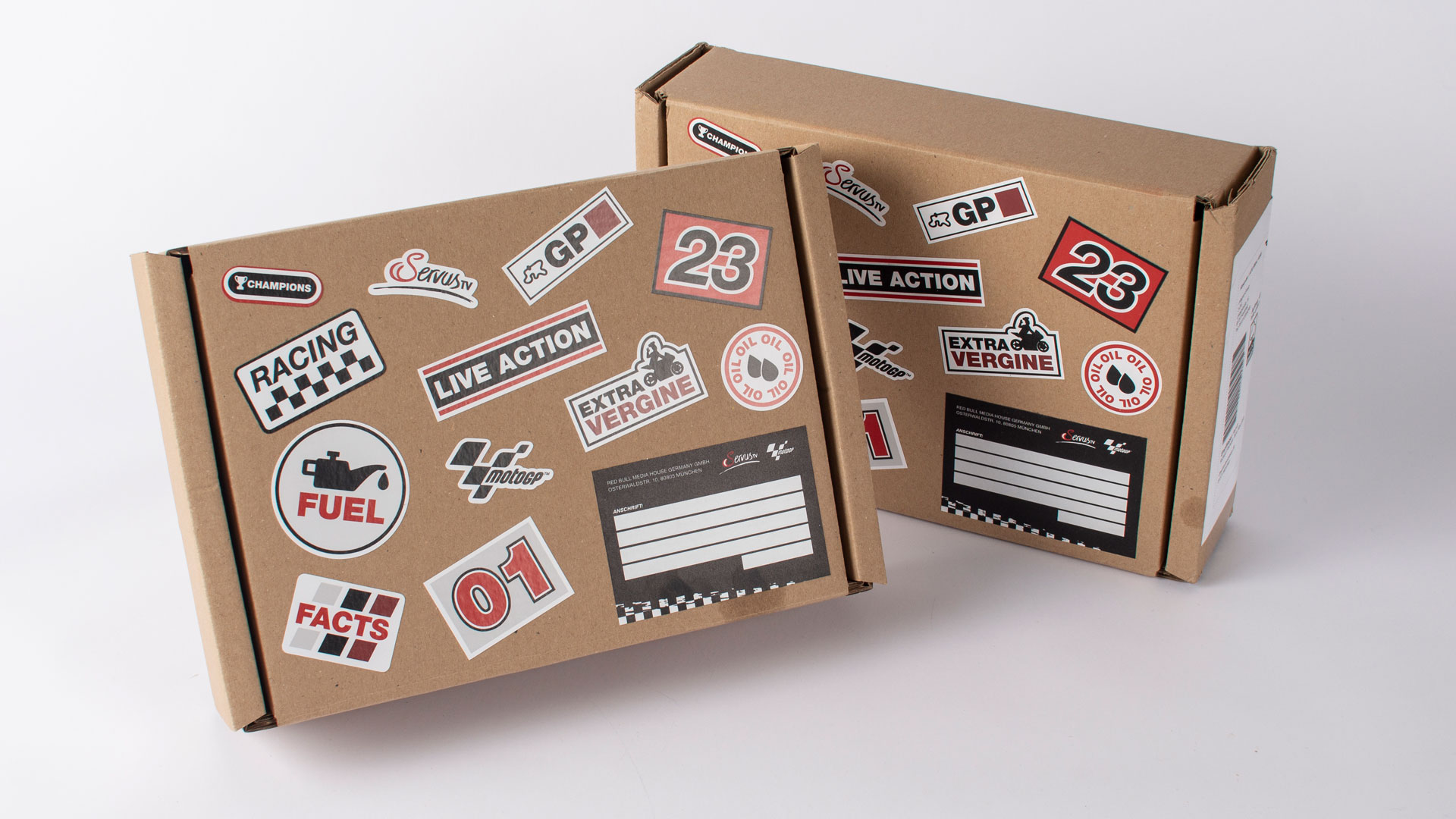 Zwei Kartons aneinander gelehnt mit Stickern darauf mit Aufschriften wie "Racing", "motoGP", "ServusTV", "Champions", "ExtraVirgine".