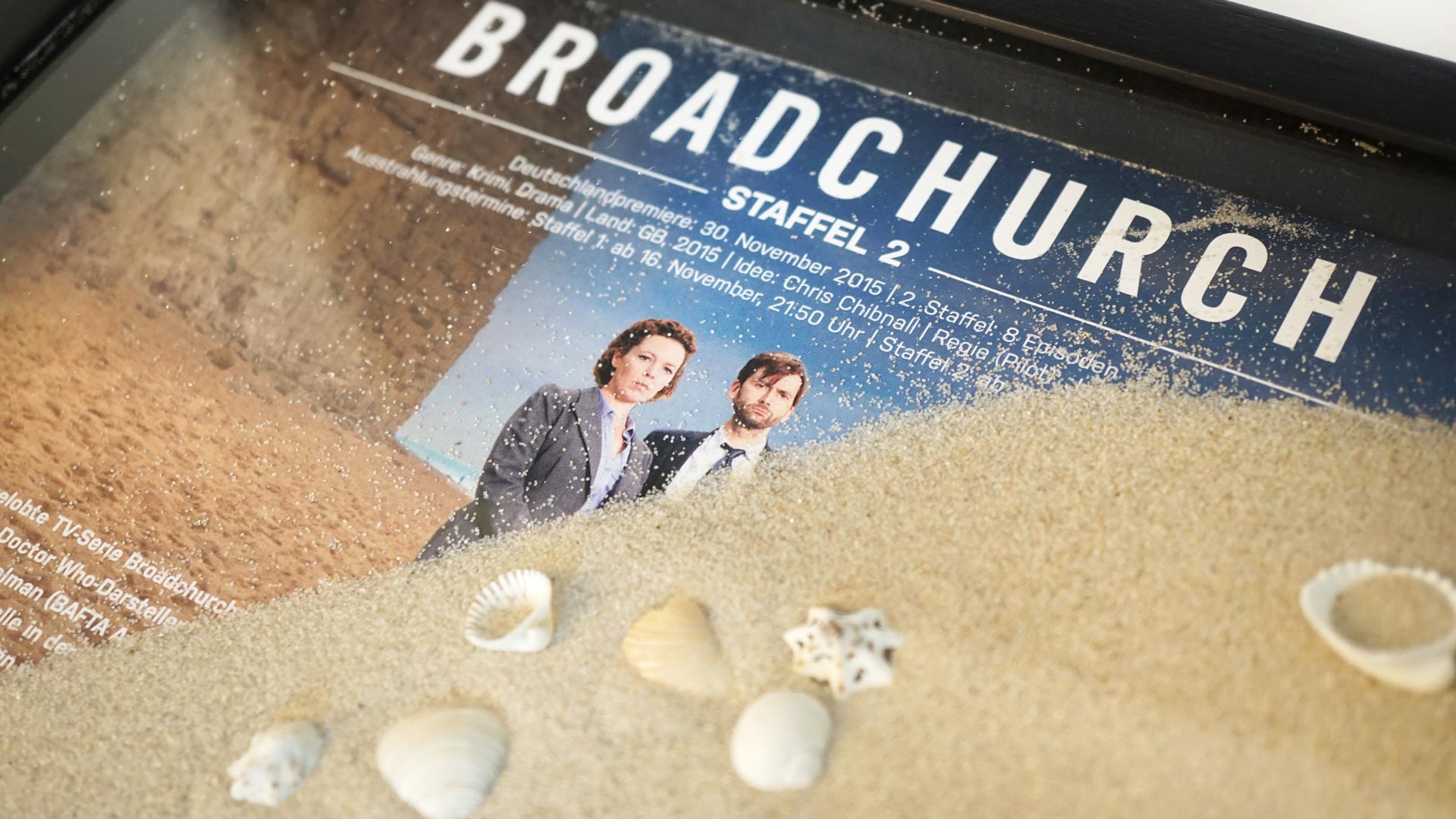 Detailaufnahme der gerahmte Pressemappe mit Sand und Motiv zur Serie Broadchurch – visuell an Strandmord angepasst.