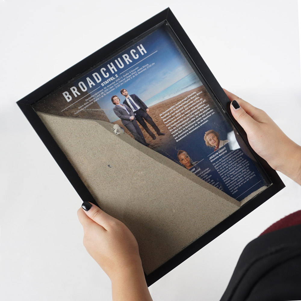 Fotorahmen wird in Händen gehalten und nach links gekippt, der Sand in dem Rahmen überdeckt das Serienplakat leicht.
