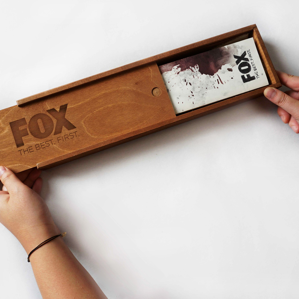 Holzschatulle mit FOX-Logo und eingraviertem Schriftzug „THE BEST. FIRST.”, in Händen gehalten, darin ein blutverschmierter Infokarton zur Serie.