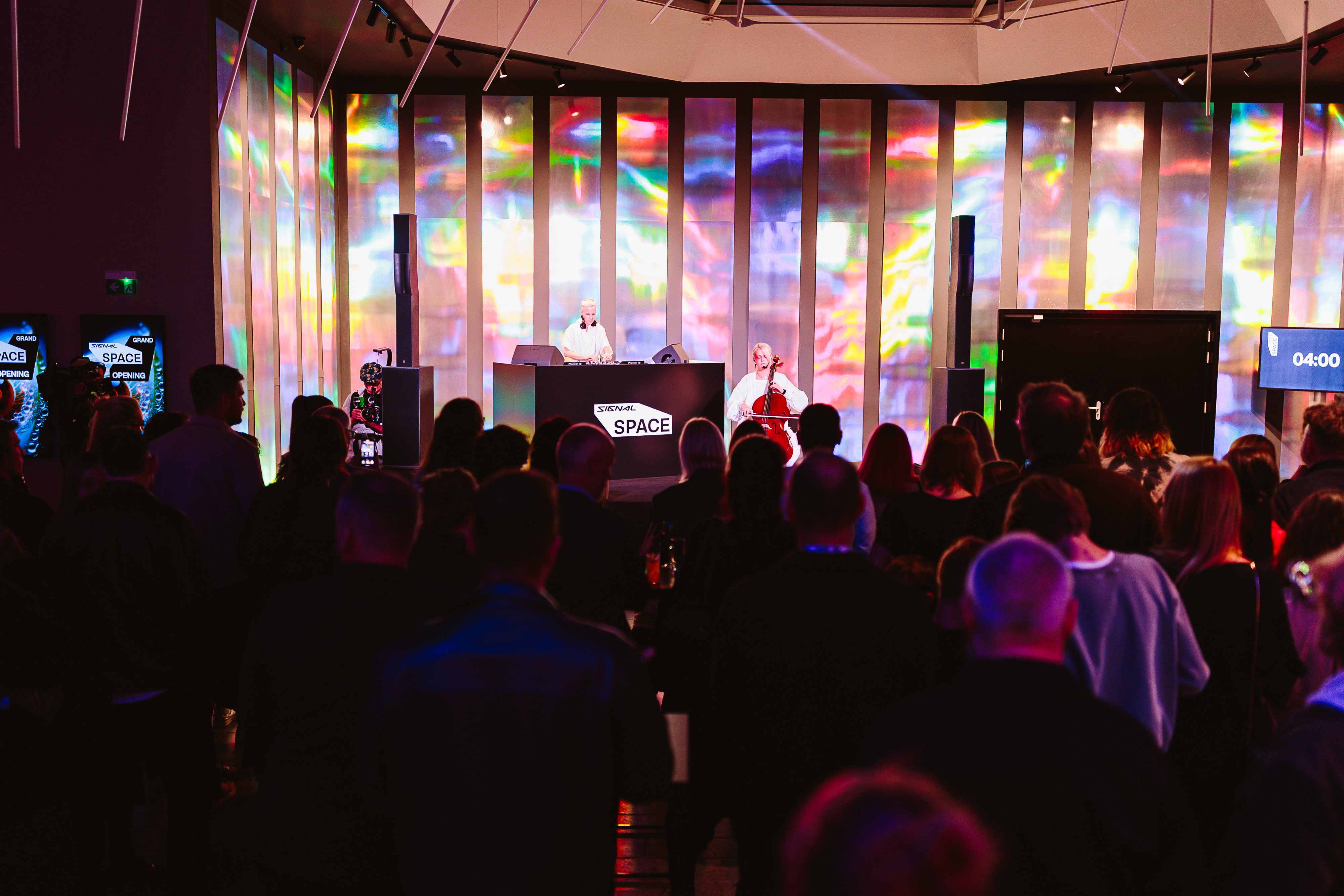 Crowd watching a live DJ and a cellist performing on stage with colorful illuminated panels behind them.