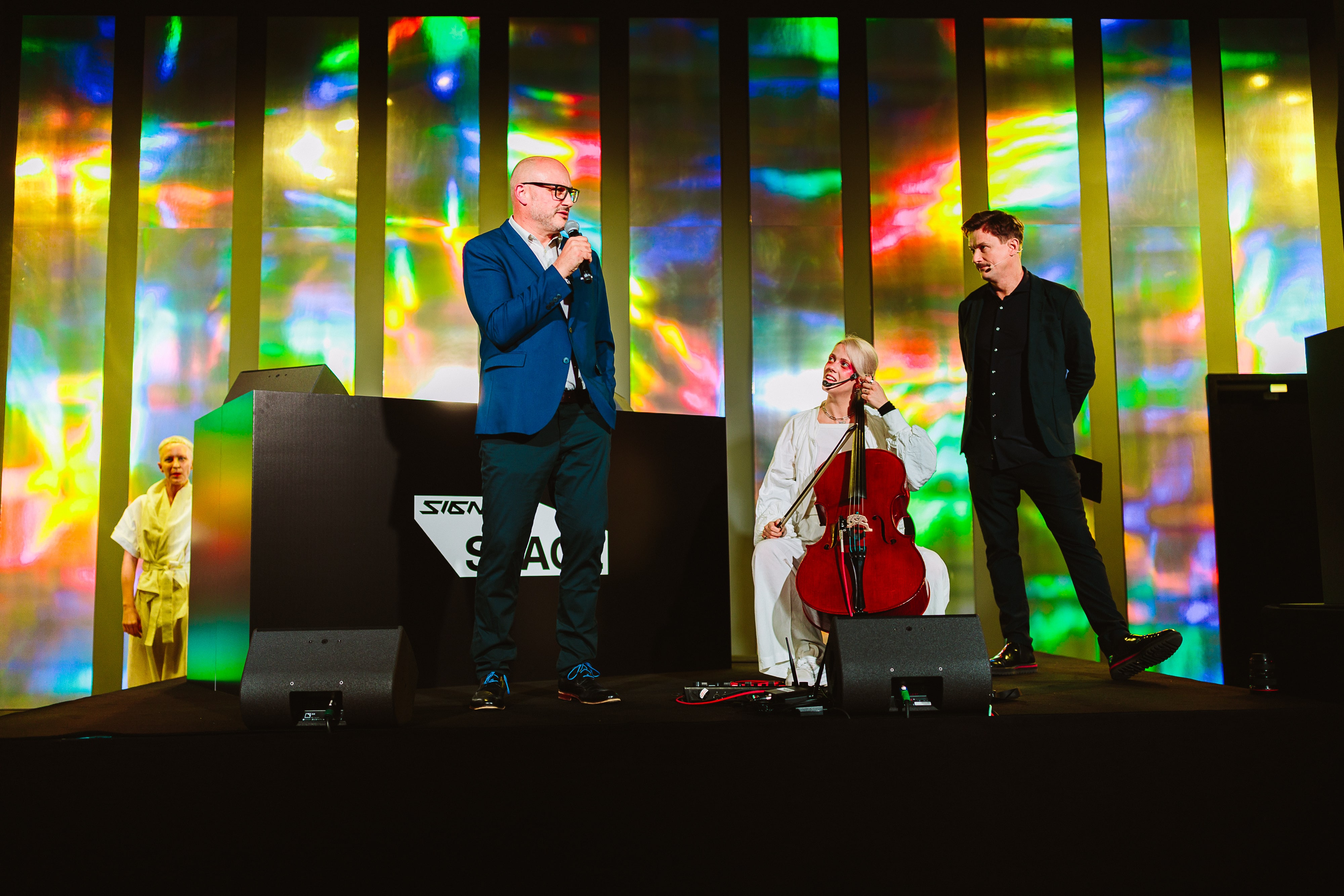 Three people on stage with colorful abstract background; a man in a blue blazer speaks into a microphone, a woman in white holds a cello, and a man in black stands nearby.