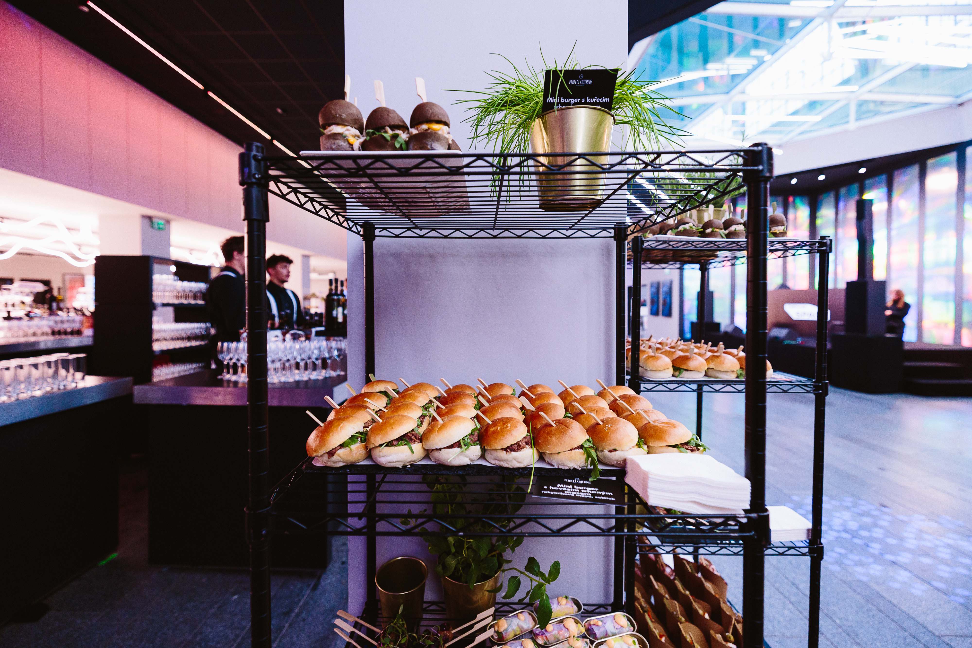 Metal rack display with rows of mini sandwiches, black mini burgers, fresh herbs, and spring rolls in a modern event space.