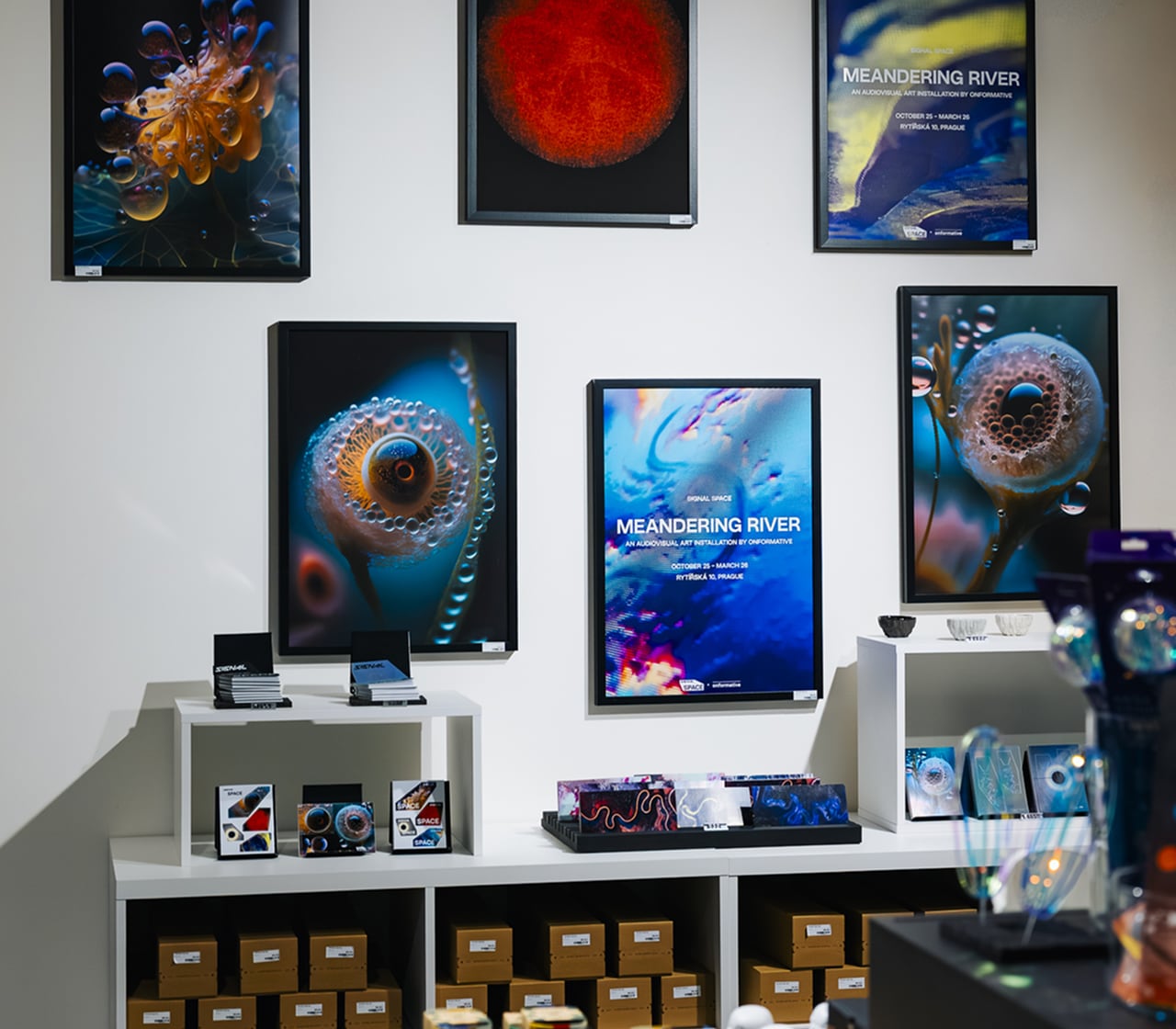 Gallery wall with framed abstract space-themed artwork and promotional posters titled 'Meandering River', with shelves displaying themed merchandise and boxes underneath.