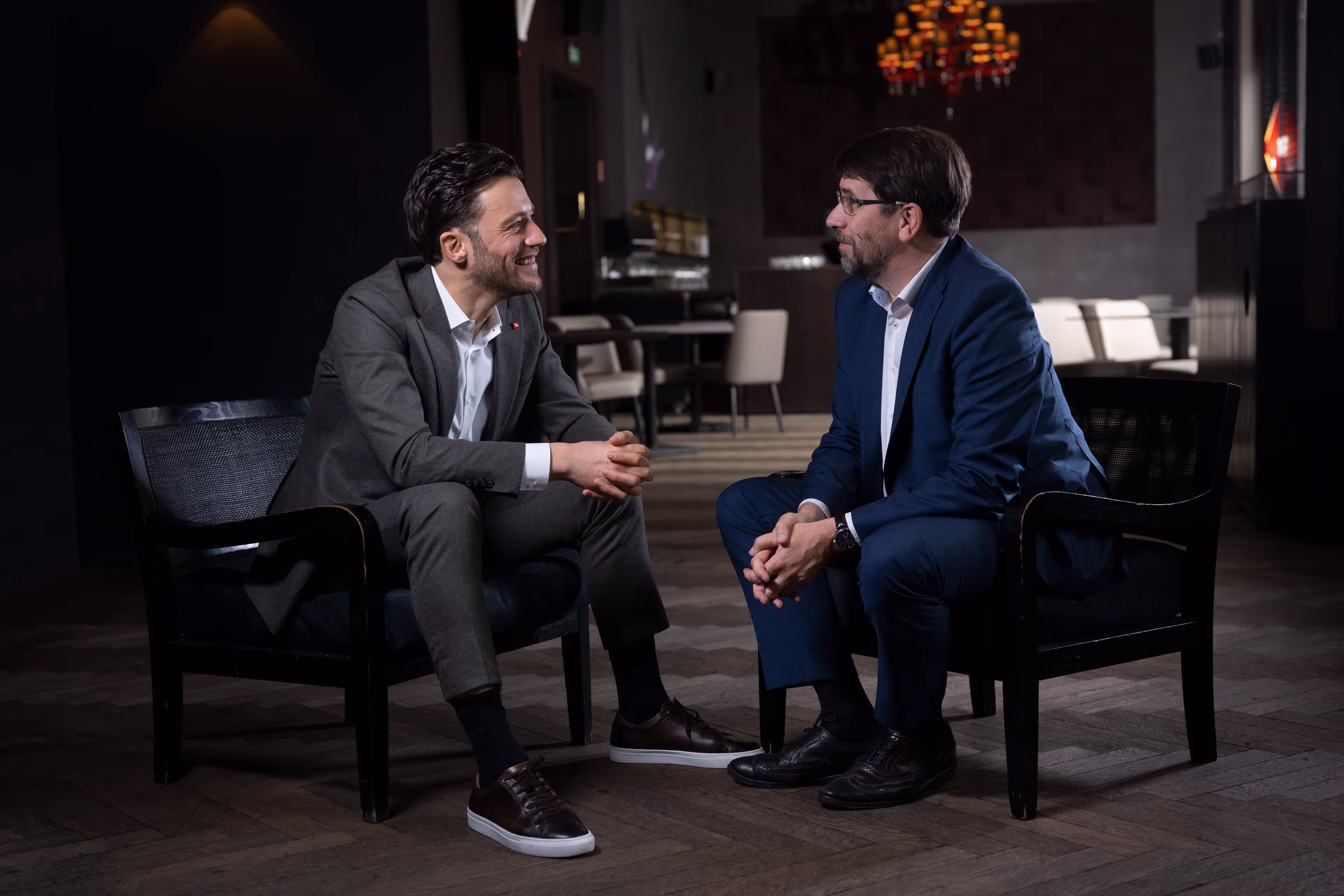 Zwei Männer im Anzug sitzen in Sesseln im Gespräch in einem stilvollen Innenraum.