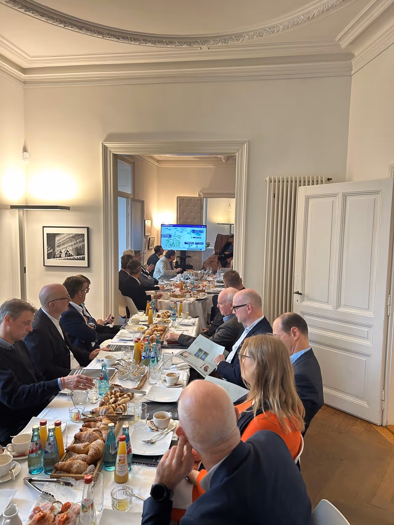 Menschen in formeller Kleidung sitzen an einem langen gedeckten Tisch mit Croissants, Getränken und Zeitungen in einem elegant eingerichteten Raum.