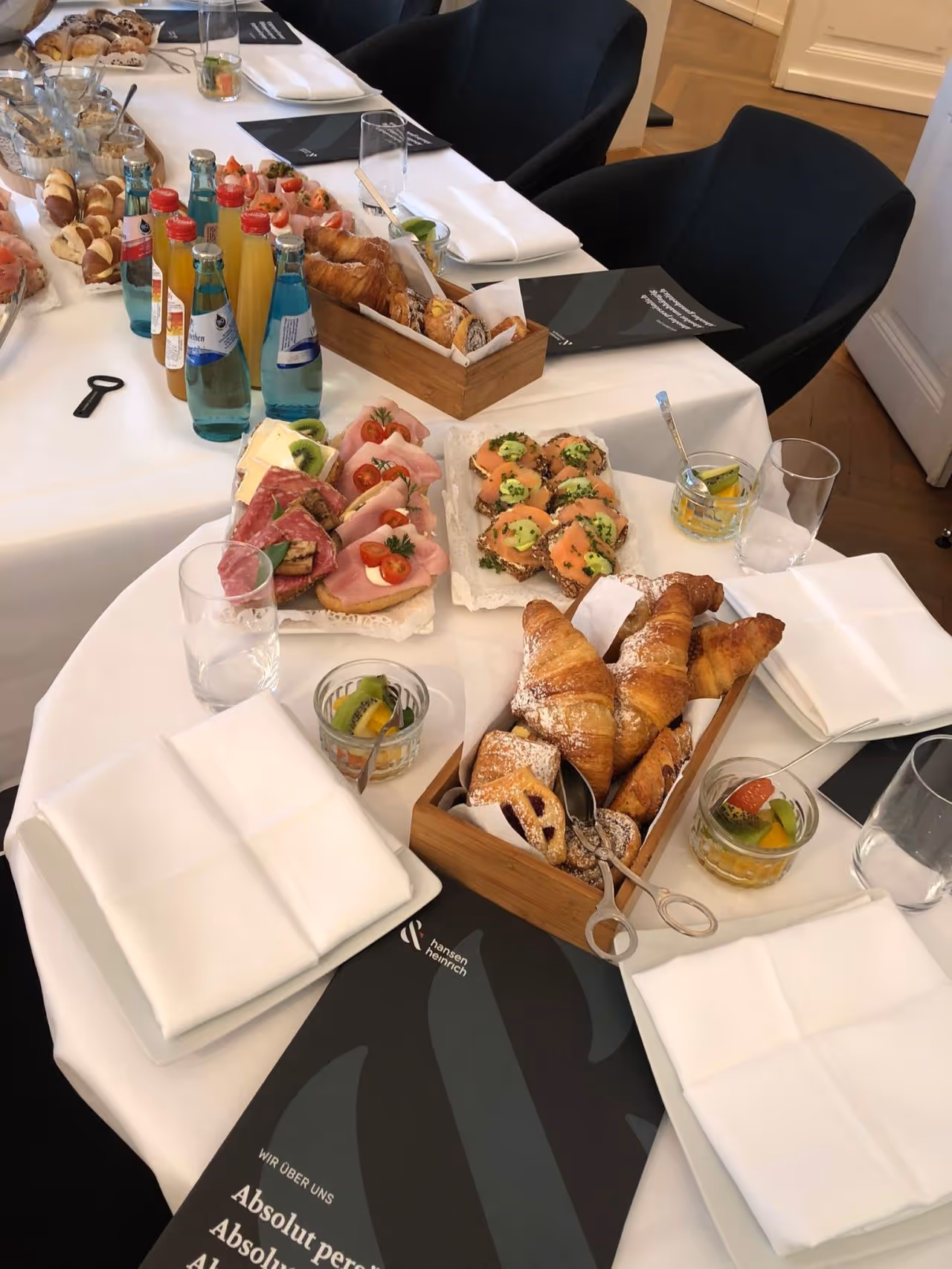 Ein gedeckter Tisch mit verschiedenen Frühstücksgebäcken, belegten Broten mit Schinken und Lachs, kleinen Obstgläschen und Flaschen mit verschiedenen Getränken.