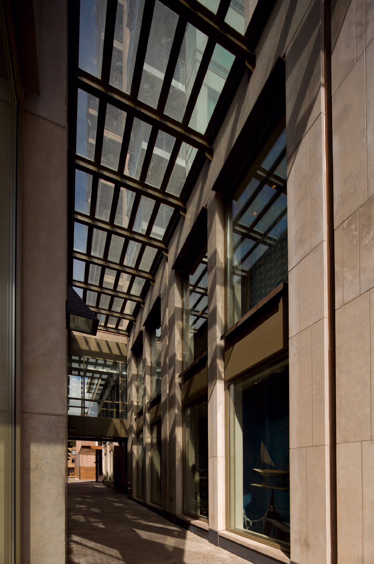Exterior corridor with a glass canopy structure casting shadows along the stone-clad wall.