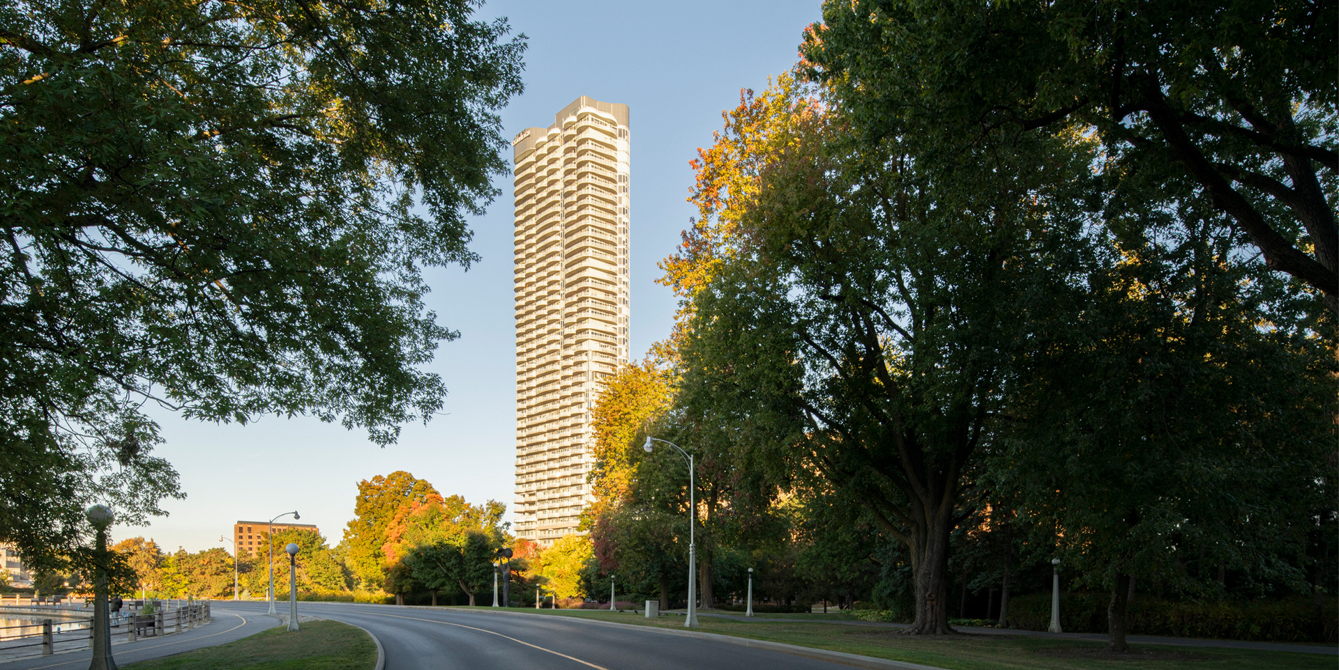 Curved road lined with street lamps and large trees with a tall multi-unit residential building in the background against a clear blue sky.