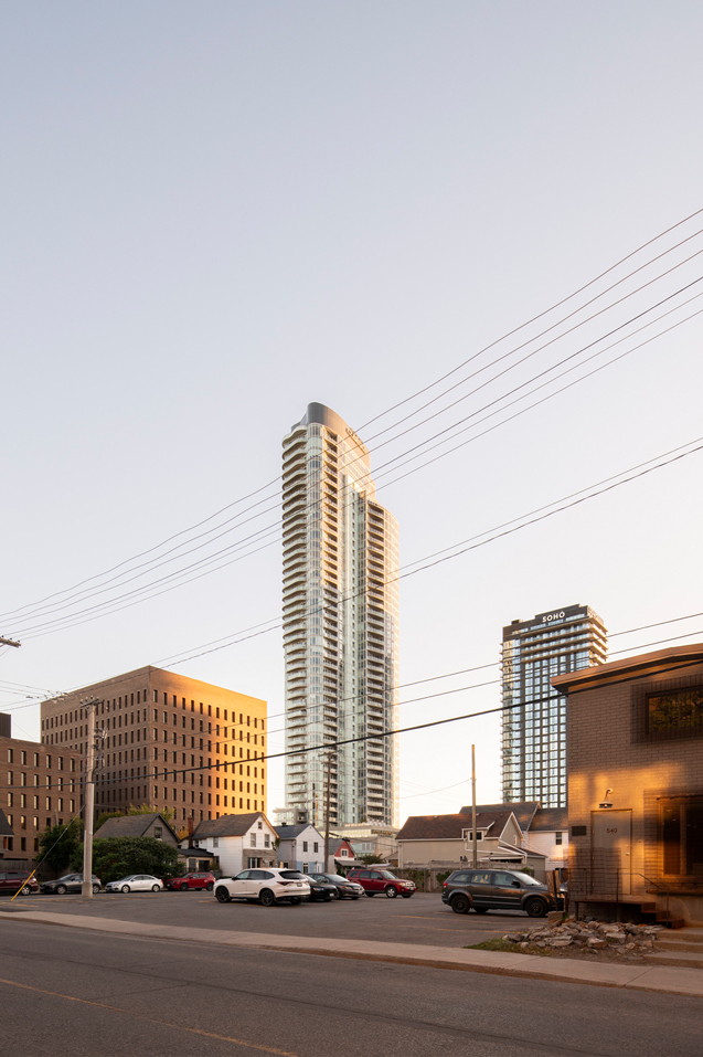 Tall modern residential skyscraper with curved balconies illuminated by sunset, surrounded by smaller buildings and parked cars in a parking lot.
