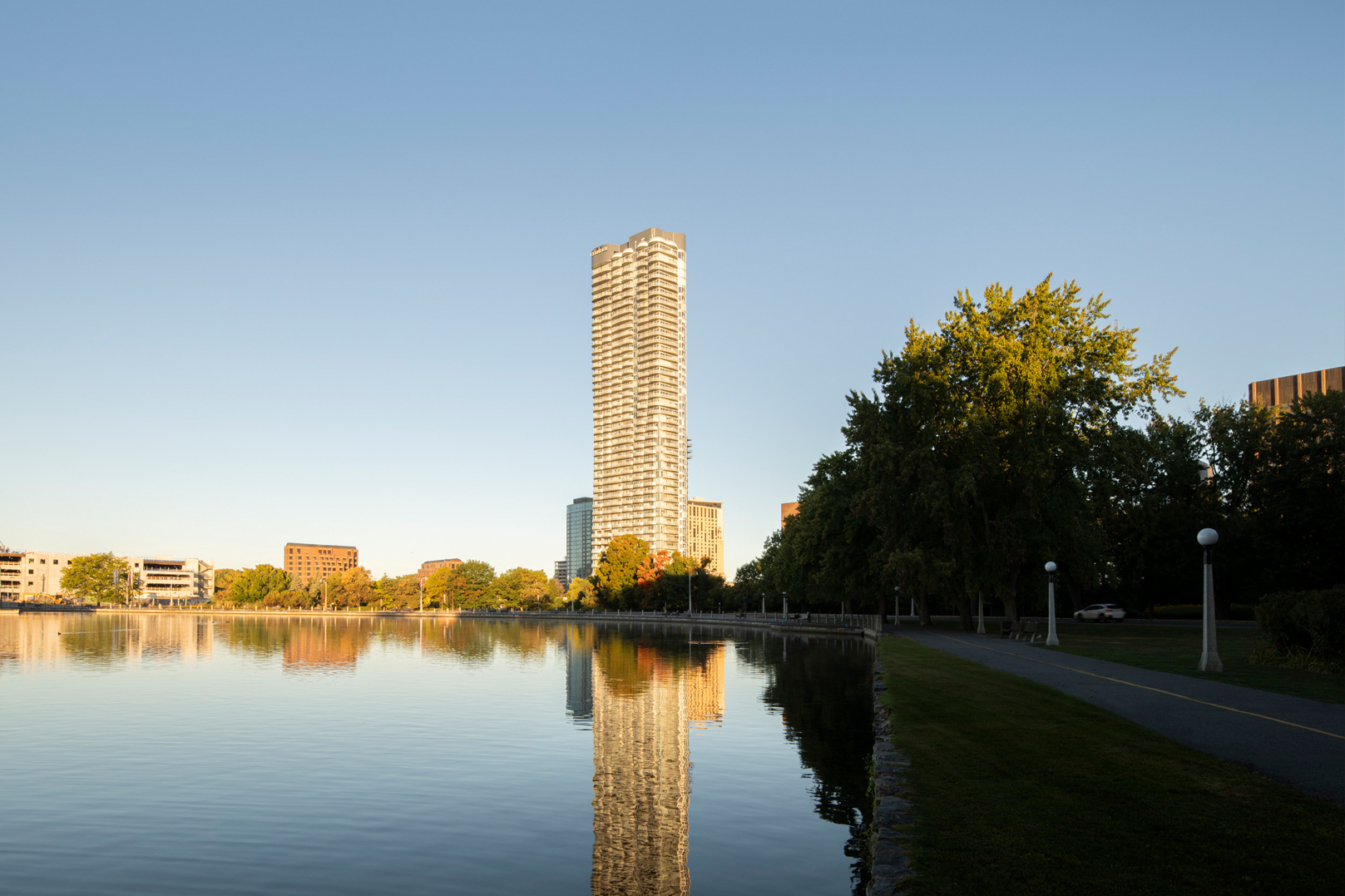 Tall modern multi-storey residential building reflecting in a lake near a tree-lined pathway at sunset.