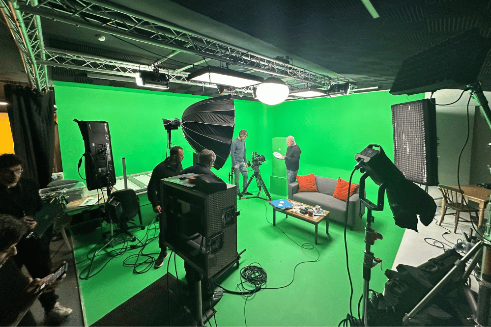 Film crew working on a green screen set with camera, lights, sofa, and props.