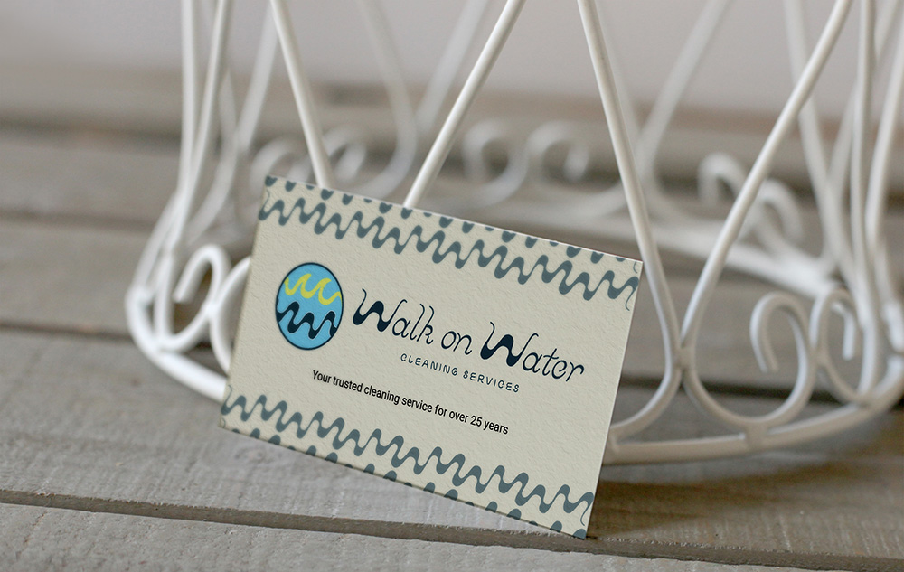 Business card for Walk Water Cleaning Services LLC with a blue and green circular logo, placed on a wooden surface in front of a white decorative metal object.