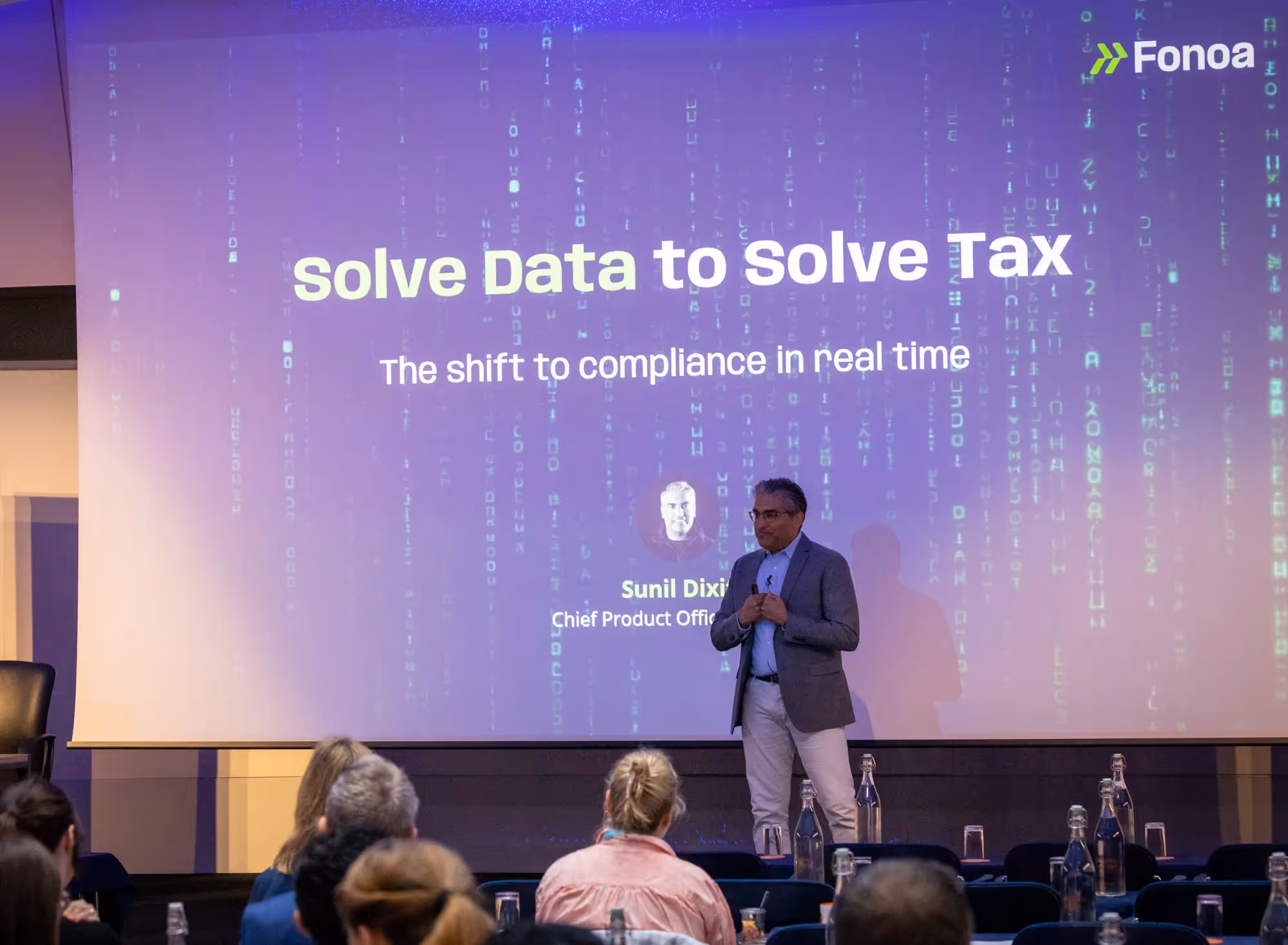 Sunil Dixit, Chief Product Officer, Fonoa, on stage at 7th Annual VAT Management Summit in London. Behind him is a large projected slide with the title “Solve Data to Solve Tax” in bold white and green text. Below that, the subtitle reads “The shift to compliance in real time.” The background of the slide displays a digital matrix-style pattern of falling green code. The audience is seated in the foreground, facing the speaker.