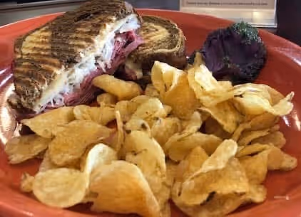 Grilled sandwich with layers of meat and melted cheese served with potato chips on an orange plate.