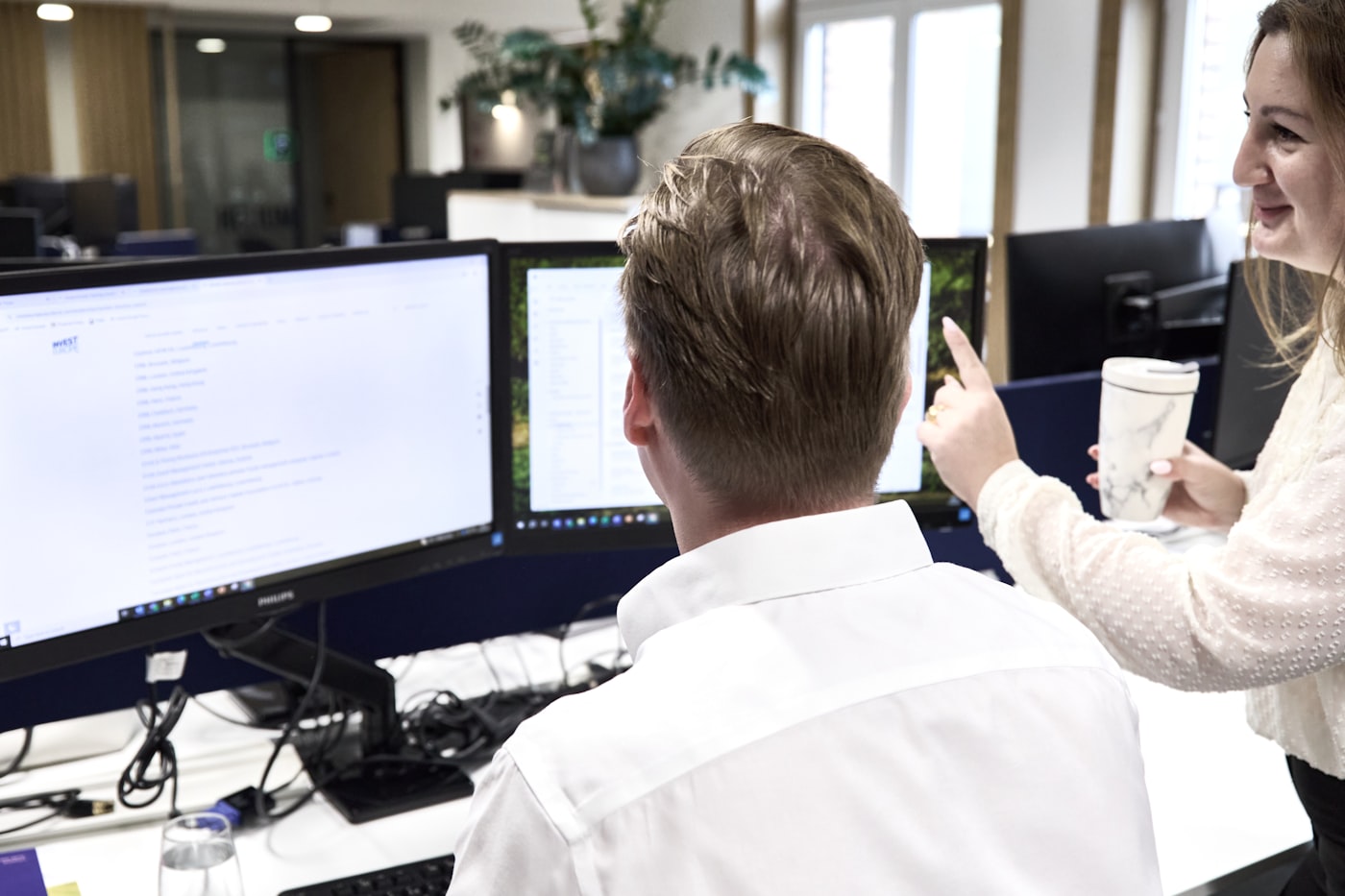 An IT specialist points at a dual-monitor workstation while explaining something to a colleague in a bright modern office