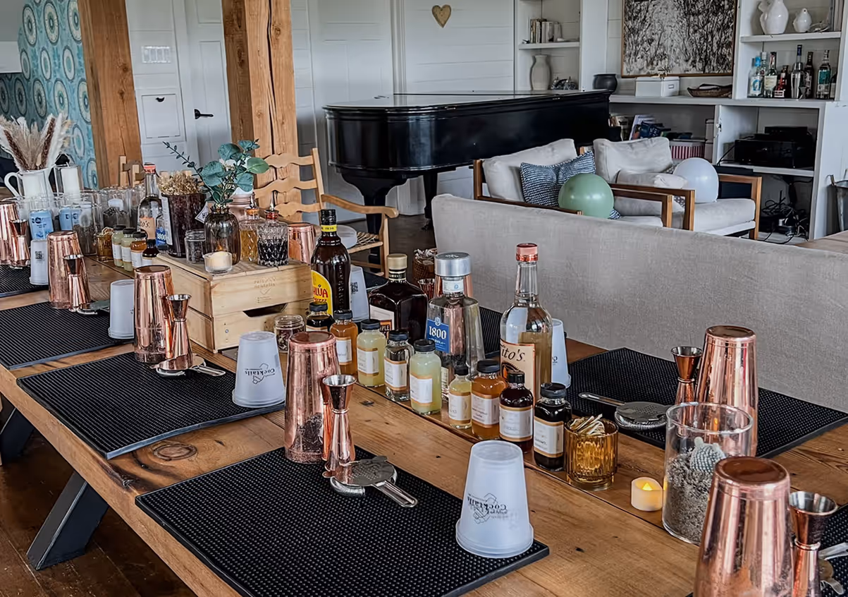 Wooden table set up for cocktail making with copper shakers, measuring jiggers, various bottles of liquor and mixers, and bar tools in a cozy living room with a black grand piano in the background.