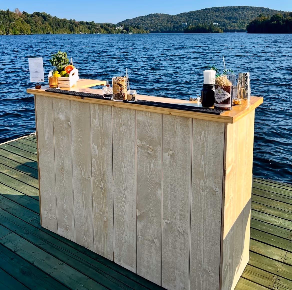 Wooden outdoor bar set up on a green dock with a lake and tree-covered hills in the background.