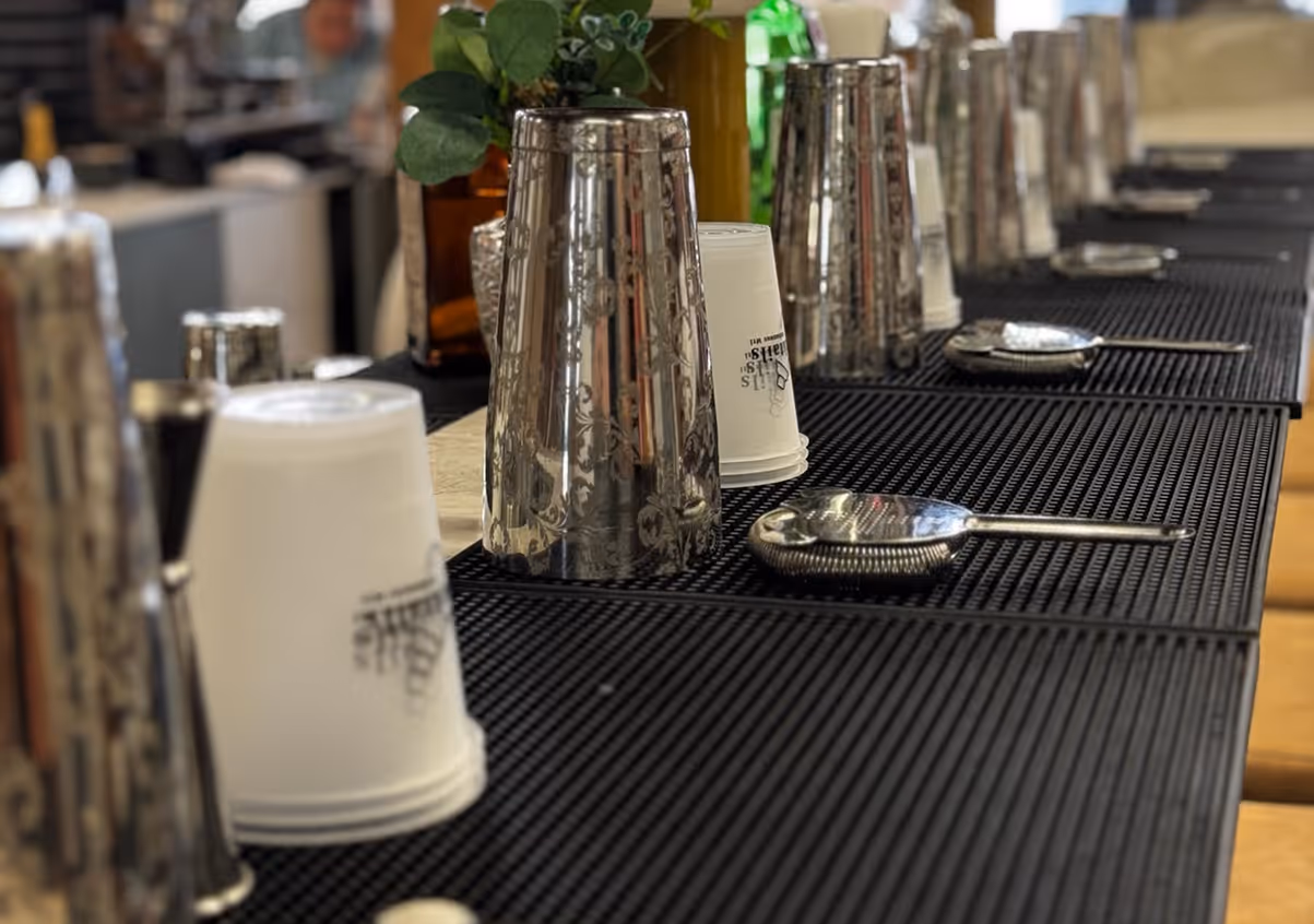 Aménagement d'un atelier de mixologie en groupe avec des tapis de bar noirs, des shakers à cocktails décorés, des gobelets en plastique réutilisables et des passoires à cocktails, le tout disposé sur un comptoir de bar.