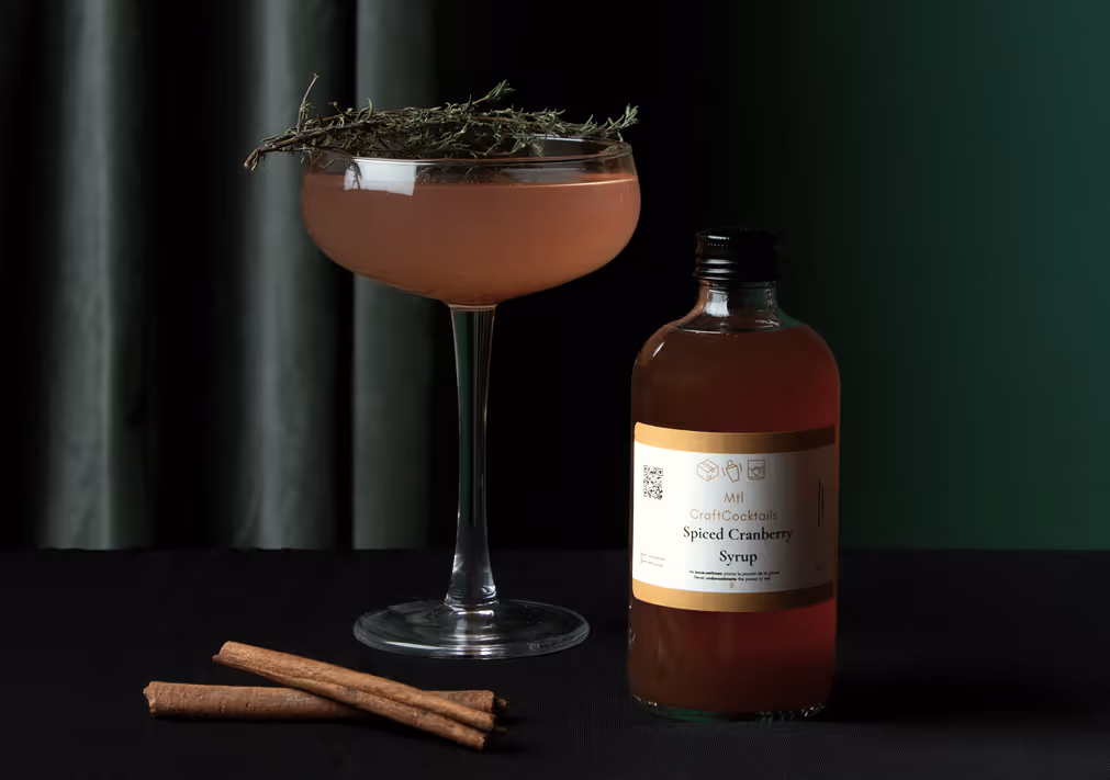 Un verre à cocktail contenant une boisson rose garnie d'un brin d'herbes aromatiques, à côté d'une bouteille de MTL Craft Cocktails étiquetée « Sirop de canneberge épicé » et de deux bâtonnets de cannelle sur une surface sombre.