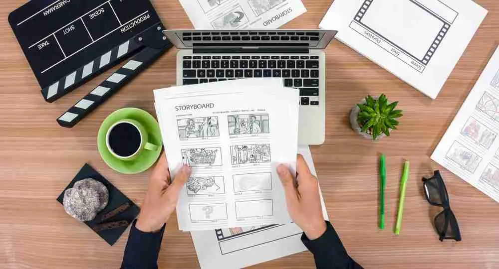 bureau avec un ordinateur un clap de cinéma une tasse de café des papiers, un story board tenu par deux mains, des stylos des lunettes de soleil