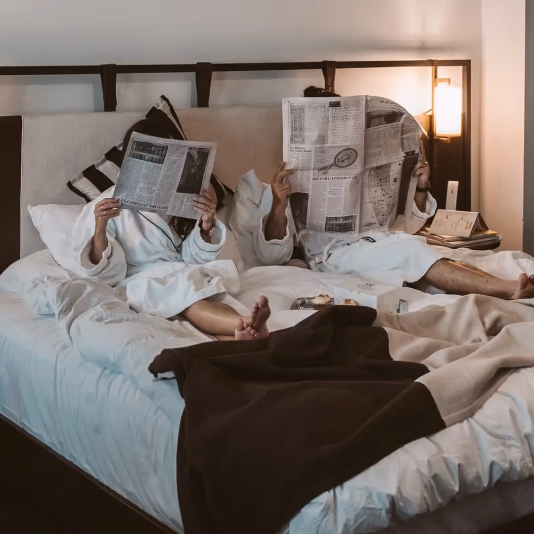 Couple in white bathrobes lying in bed, each holding a newspaper that covers their face, with breakfast on the bed.