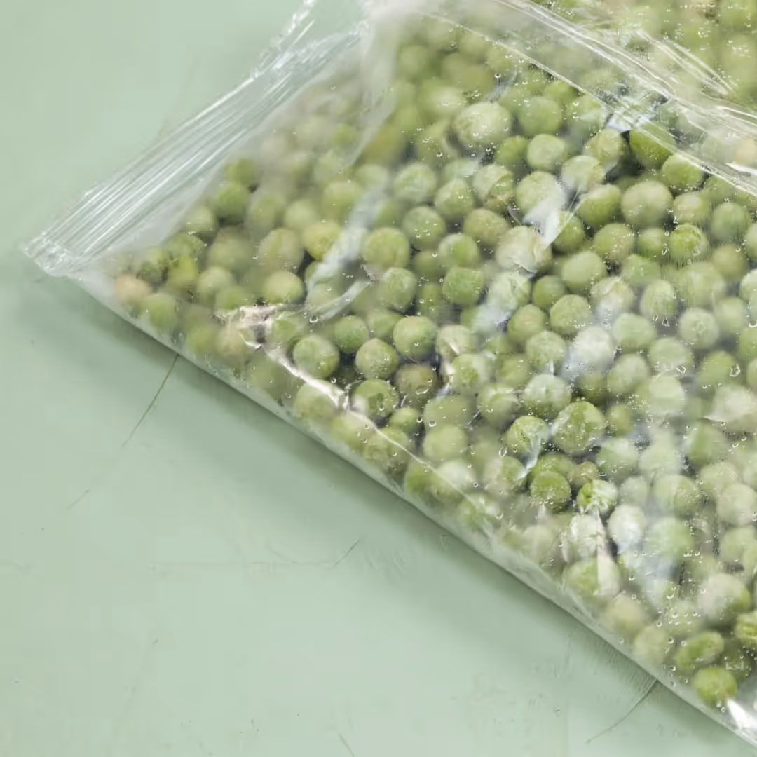 Close-up of a plastic bag filled with frozen green peas on a light green surface.
