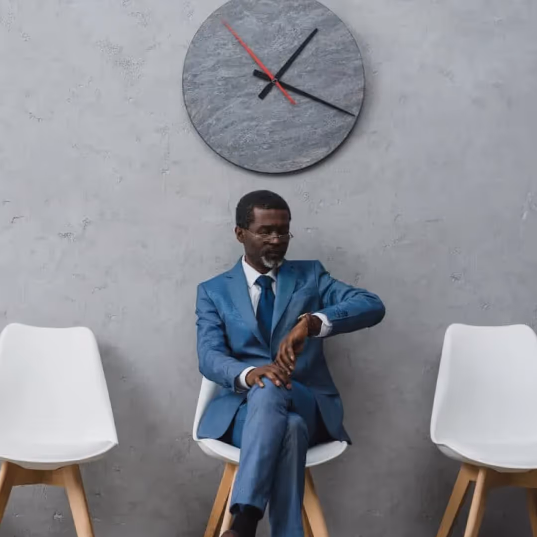 Man in a blue suit sitting between two white chairs, looking at his watch with a large wall clock above him.