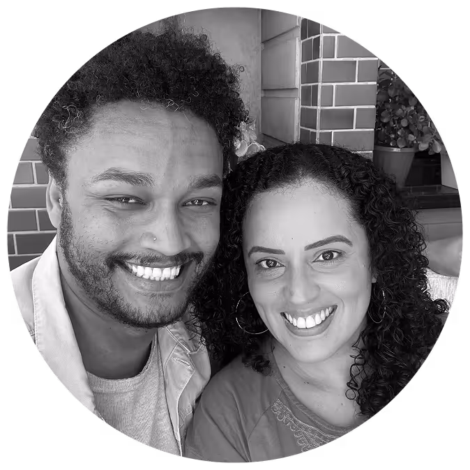 A smiling man and woman pose closely together for a selfie. The man has short curly hair, a beard, and a nose ring, while the woman has long curly hair and is wearing hoop earrings. They are sitting in front of a tiled wall with potted plants in the background. The image is black and white and cropped into a circular frame.