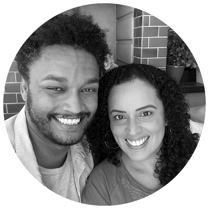 A smiling man and woman pose closely together for a selfie. The man has short curly hair, a beard, and a nose ring, while the woman has long curly hair and is wearing hoop earrings. They are sitting in front of a tiled wall with potted plants in the background. The image is black and white and cropped into a circular frame.