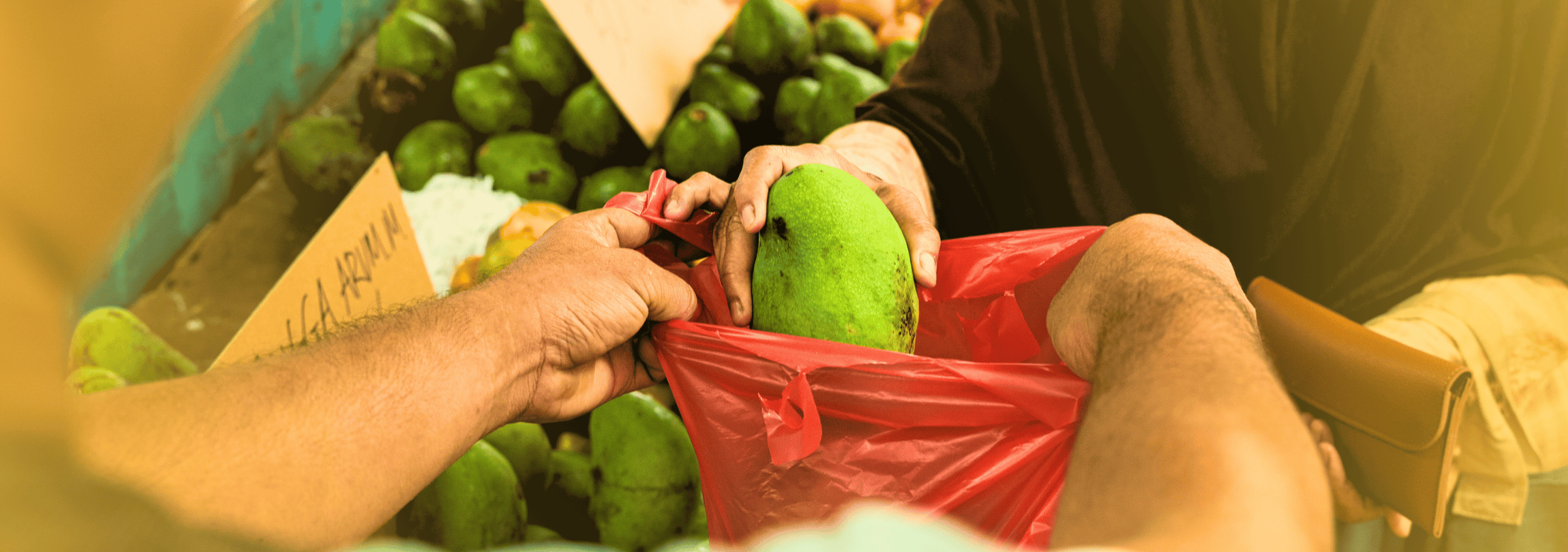 Photo of hands holding a produce bag and someone placing a mango inside