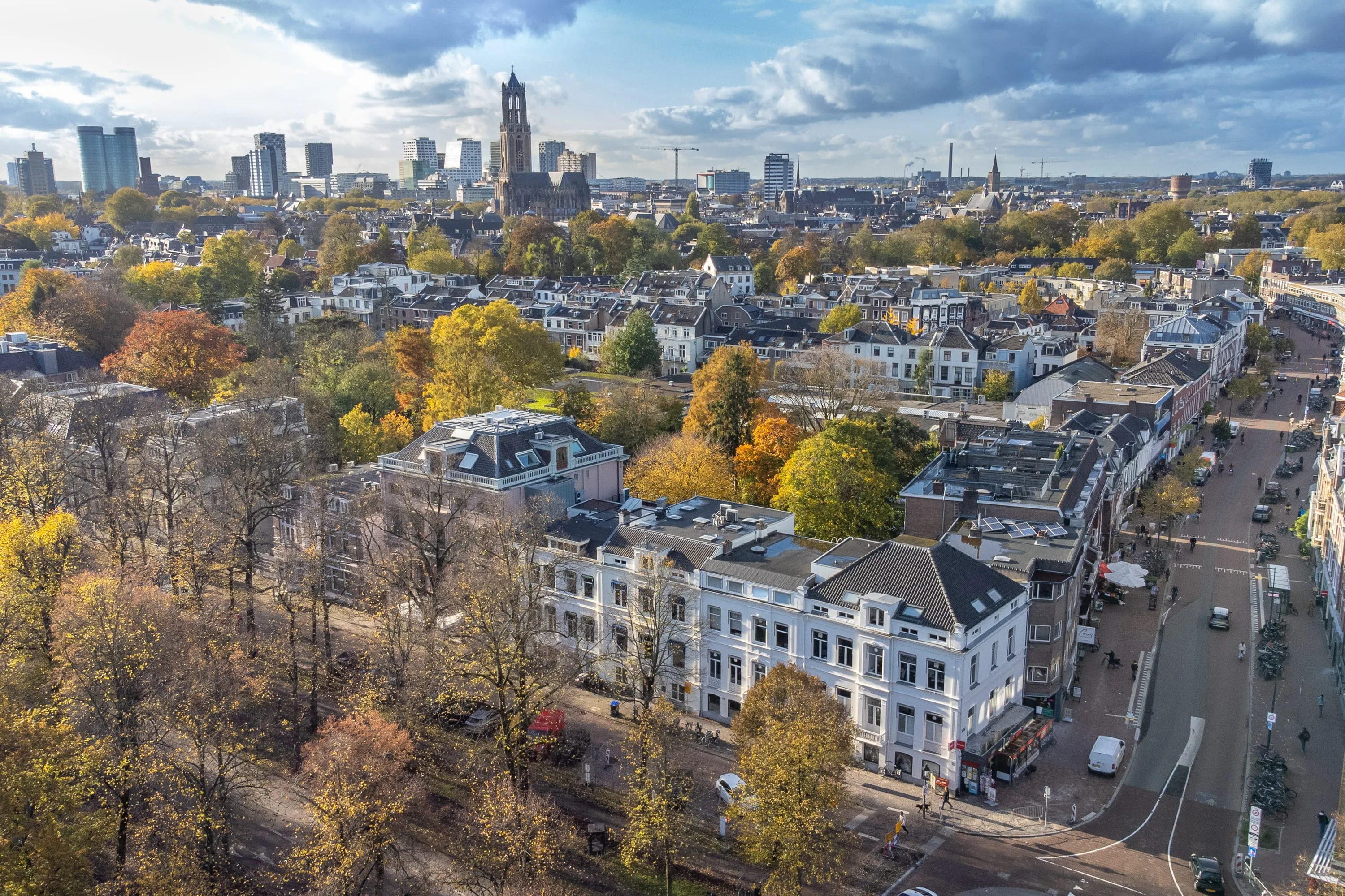 Aerial view of a cityscape in the Netherlands, featuring a mix of modern and historic buildings with trees displaying autumn colors. The skyline includes tall buildings and a tower in the background, illustrating urban life ideal for expat relocation in the Netherlands.
