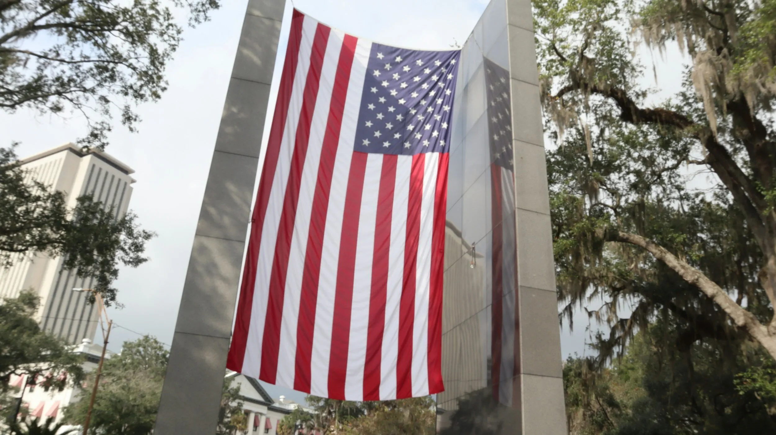 Large American flag hanging between two stone pillars with trees and buildings in the background.
