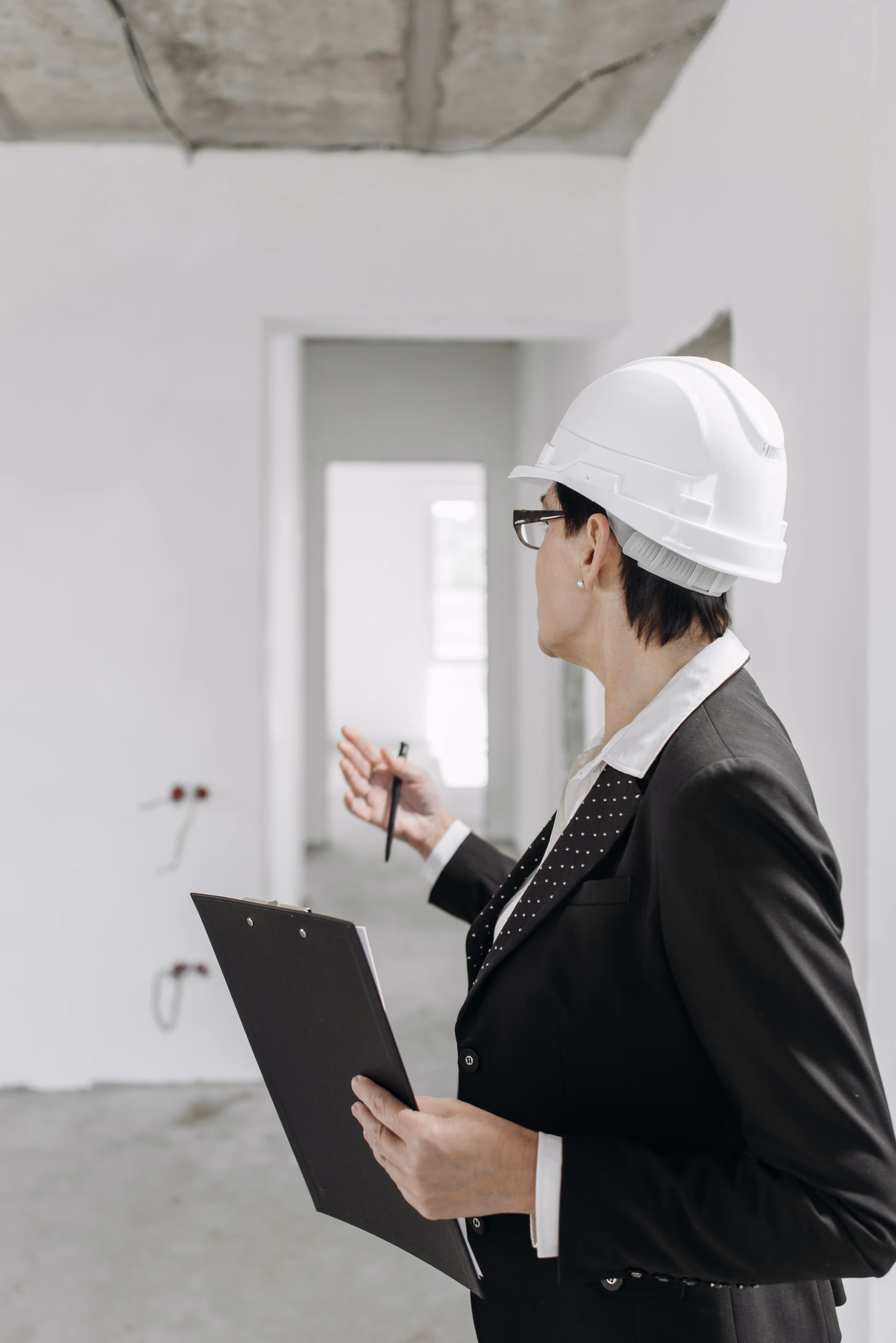 Professional woman in a black suit and white safety helmet holding a clipboard and pen inspecting an unfinished room.