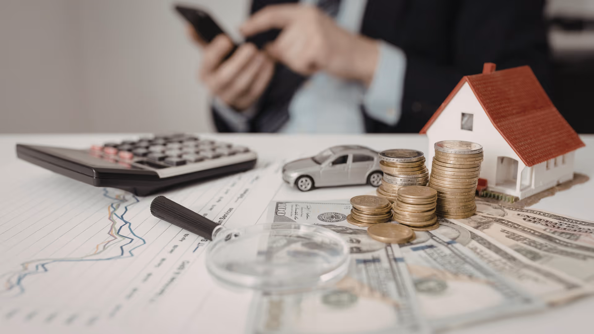 Stacks of coins, toy car, miniature house, calculator, magnifying glass, and dollar bills on a financial document with a person using a smartphone in the background.