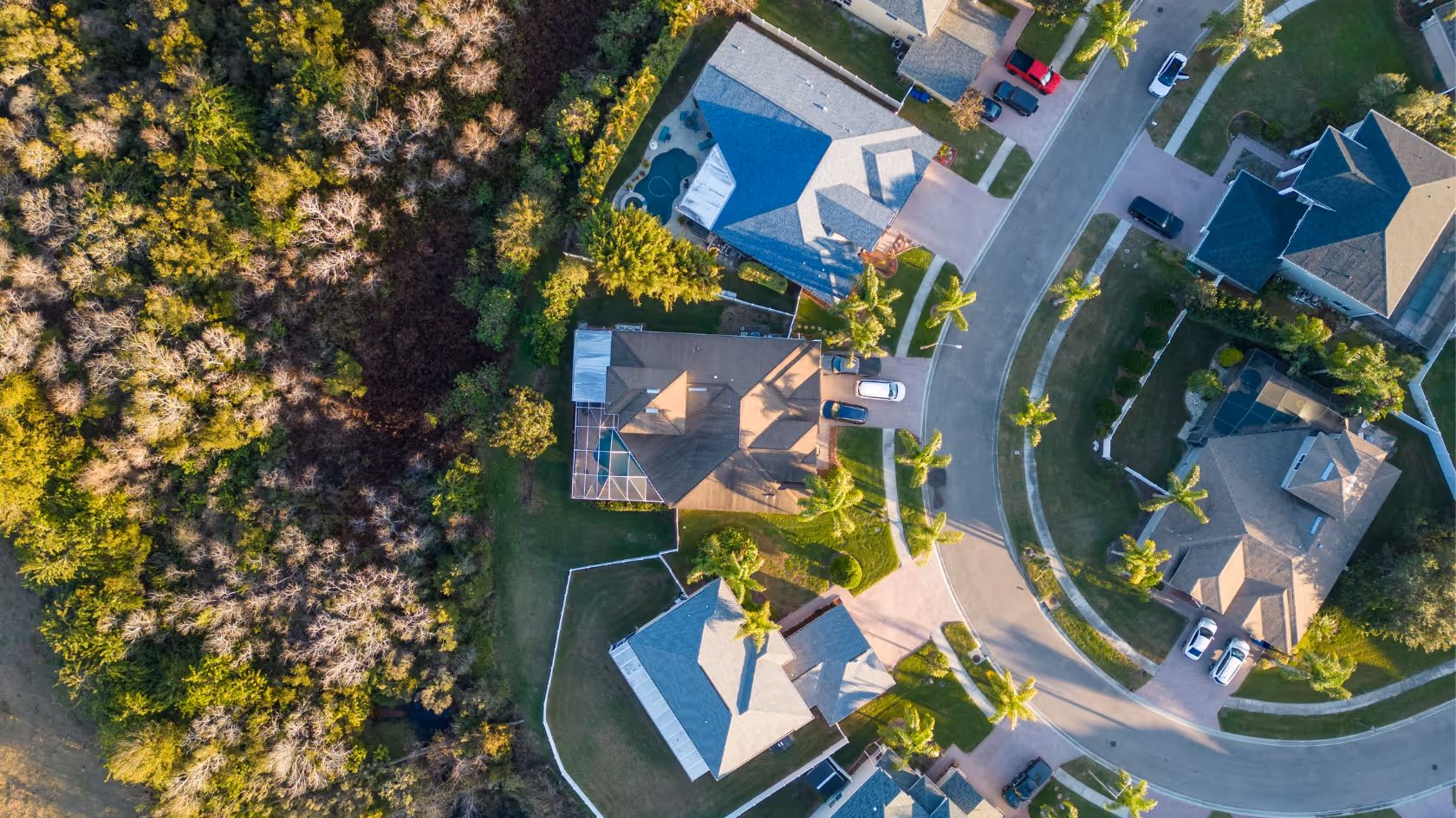 Aerial view of a suburban neighborhood with houses, curved street, cars parked in driveways, and green lawns adjacent to dense trees.