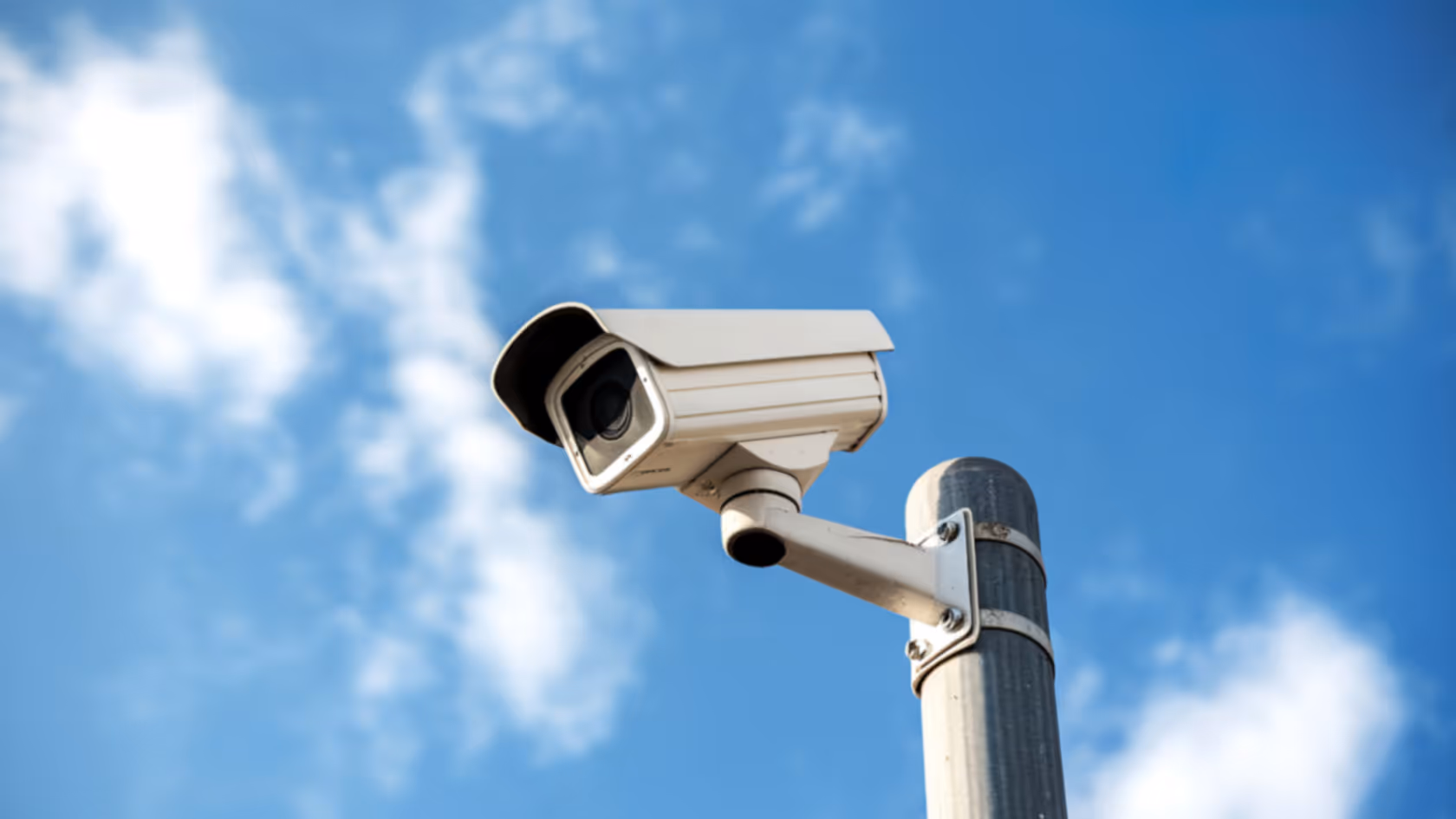 White security surveillance camera mounted on a metal pole against a blue sky with scattered clouds.