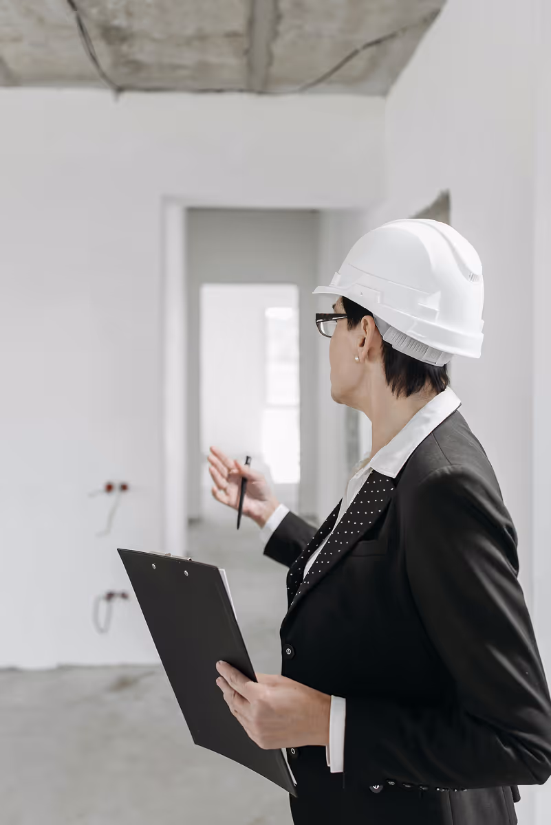 Professional woman in a black suit and white safety helmet holding a clipboard and pen inspecting an unfinished room.