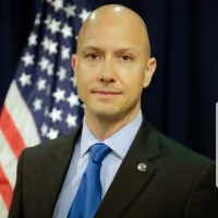 Man in suit and blue tie standing in front of an American flag and dark curtain.